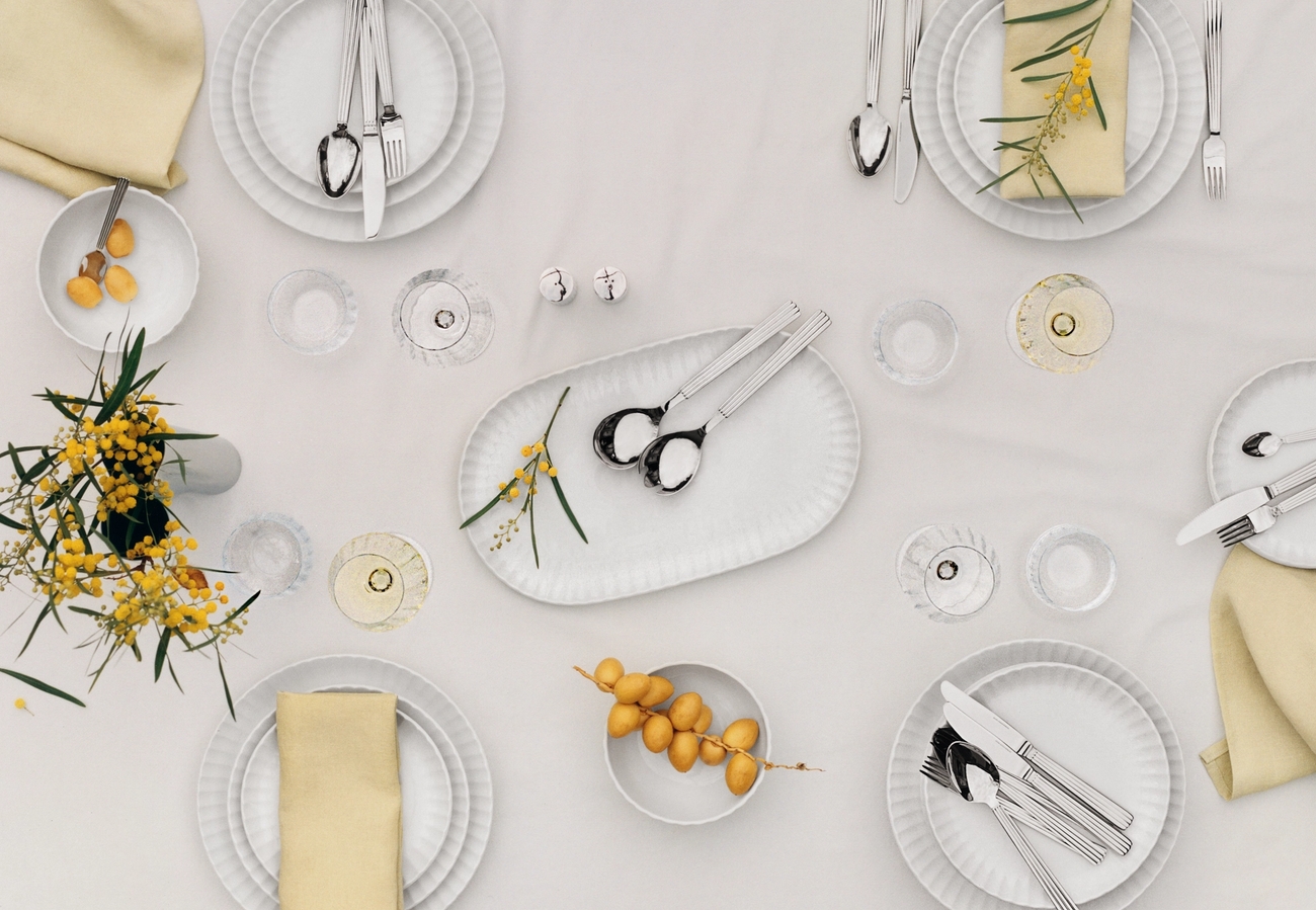 Ovanifrånvy av ett elegant matbord dukat med vita tallrikar, beige servetter, moderna bestick, vinglas och gula blommor.