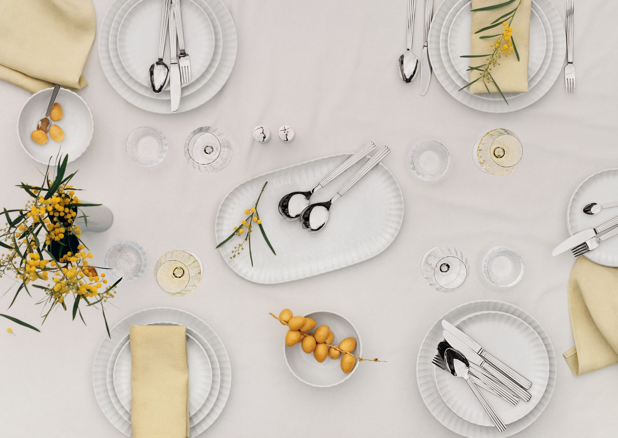 En ovanifrånvy av ett matbord dukat med vita räfflade tallrikar, silverbestick, gula servetter, mimosablommor och dadlar.