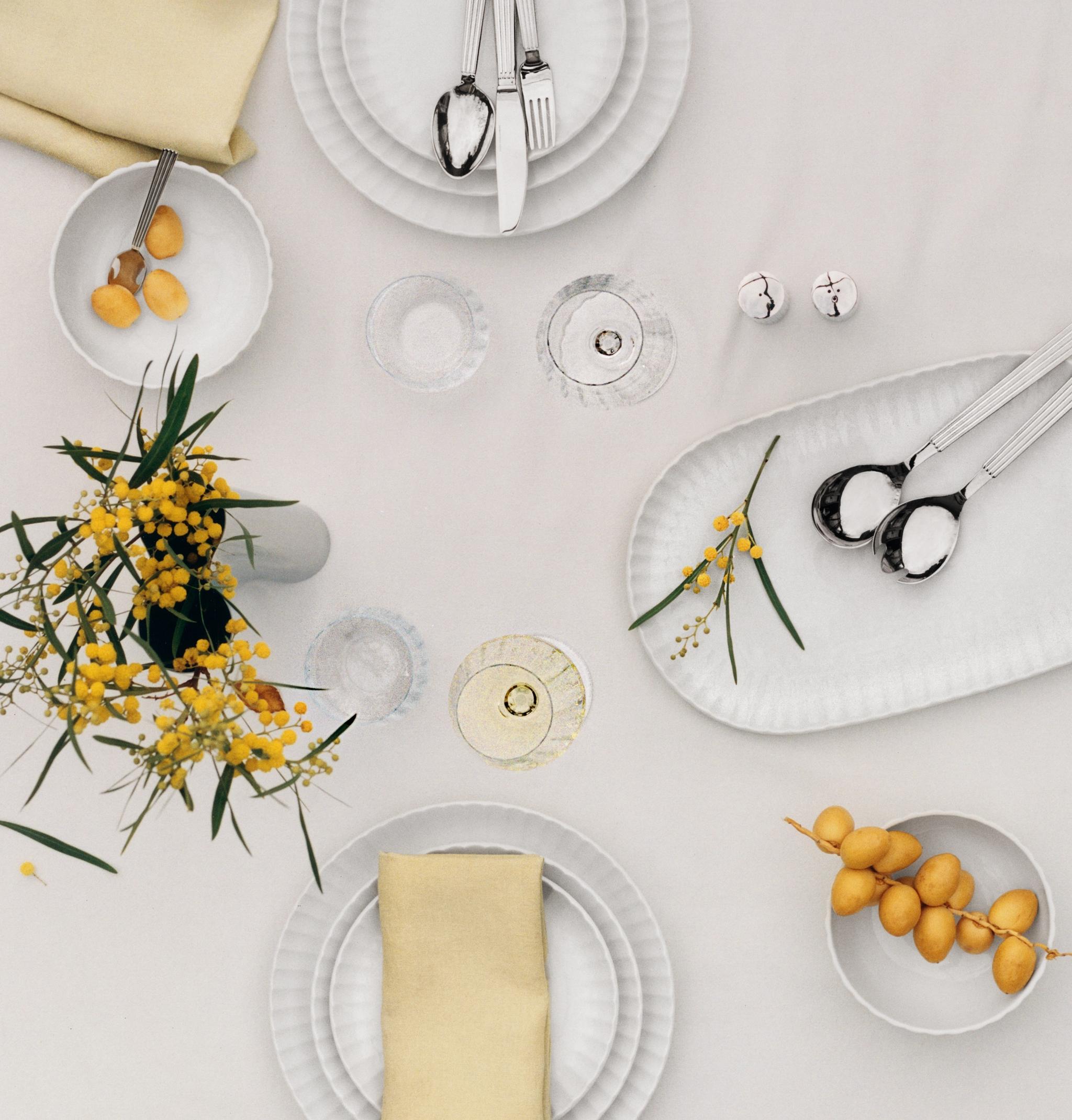 En ovanifrånvy av ett matbord dukat med vita räfflade tallrikar, silverbestick, gula servetter, mimosablommor och dadlar.