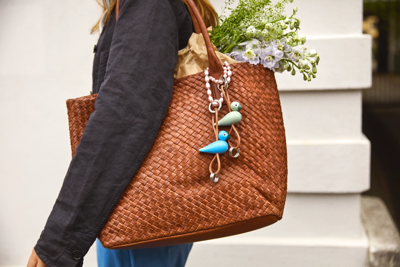 Person som bär en brun vävd läderväska med blommor och två nyckelringar med fåglar på