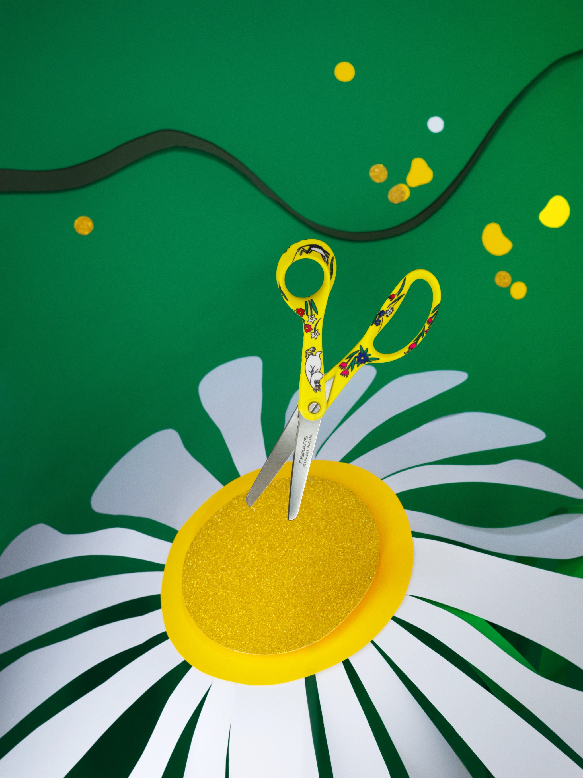 A pair of yellow patterned scissors cuts into the glittery yellow center of a large white paper daisy, against a green background with scattered confetti.