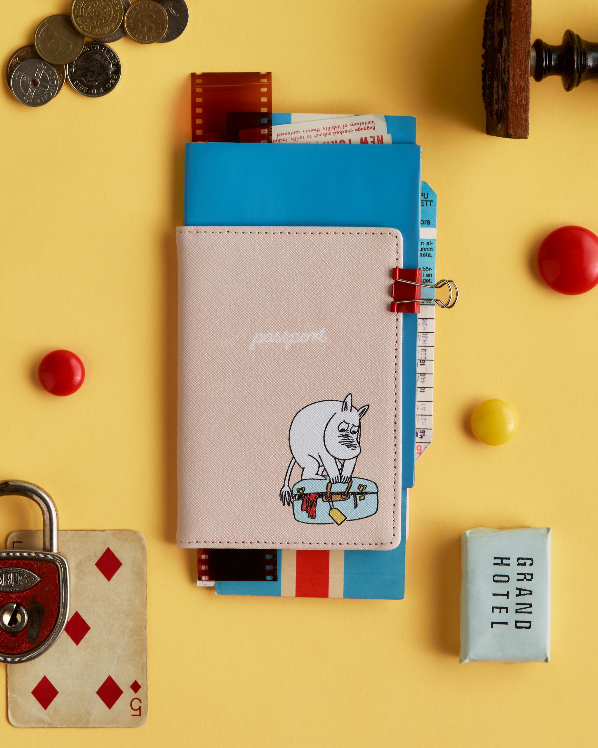 Ett ljusrosa passfodral med en Muminfigur på en resväska, omgiven av resesaker som mynt och ett hänglås på en gul yta.
