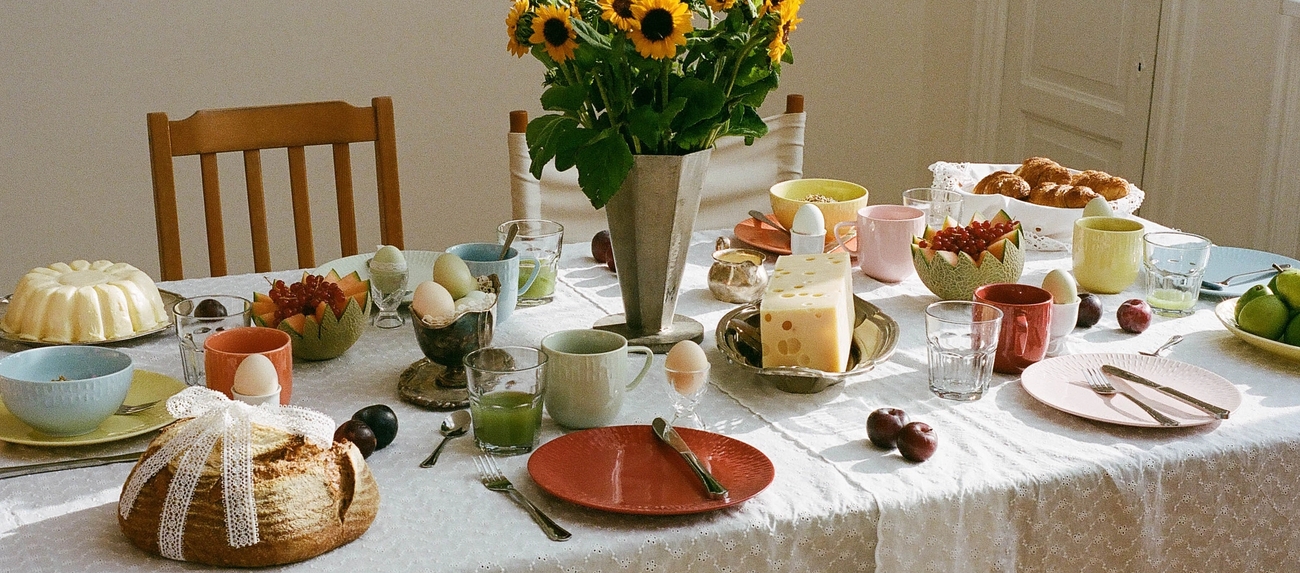 Vackert dukat frukostbord med färgglada tallrikar, bröd, ost, frukt och en bukett solrosor som mittpunkt.