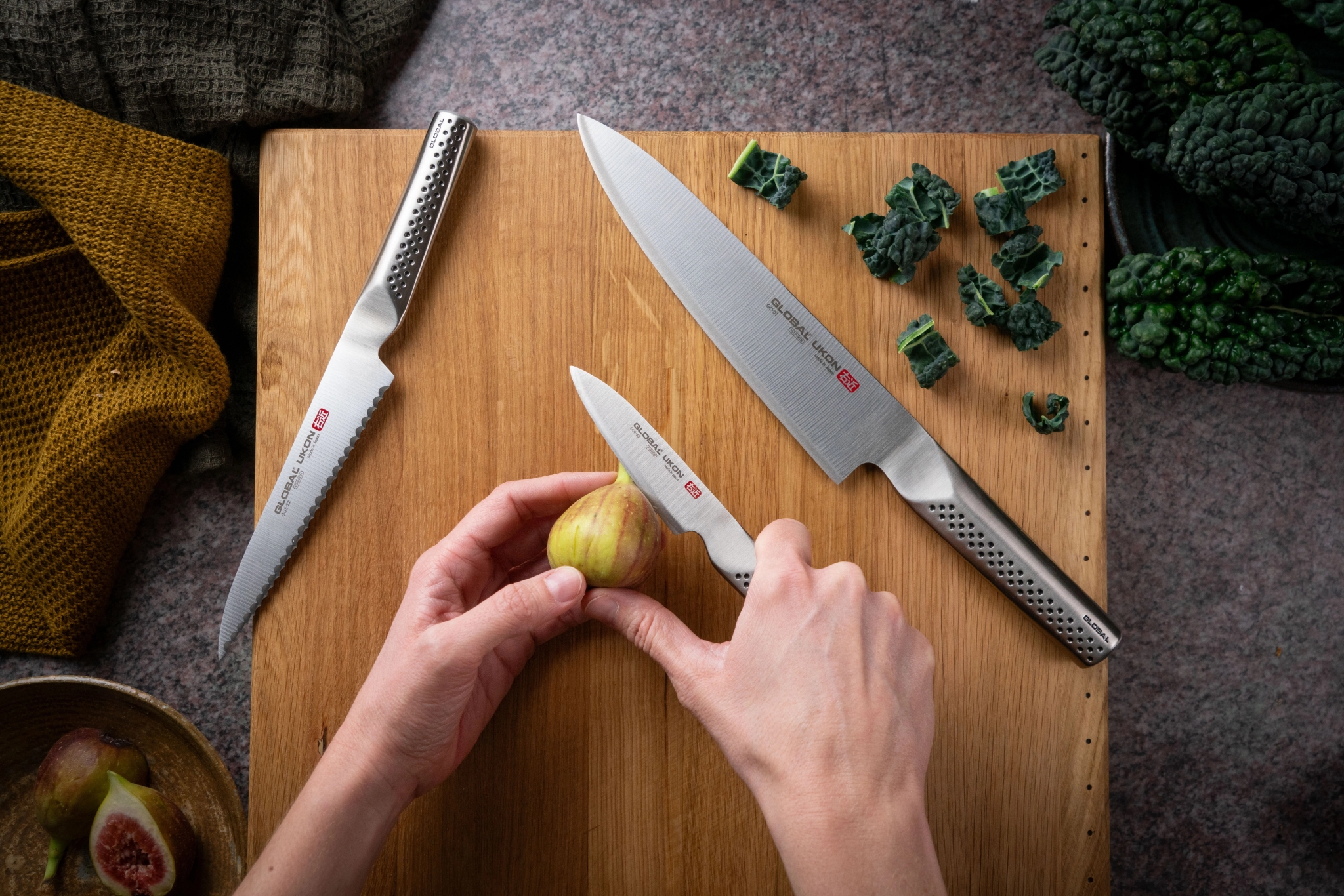Händer skär ett färskt fikon med en Global-kniv på en skärbräda omgiven av köksknivar och grönkål.