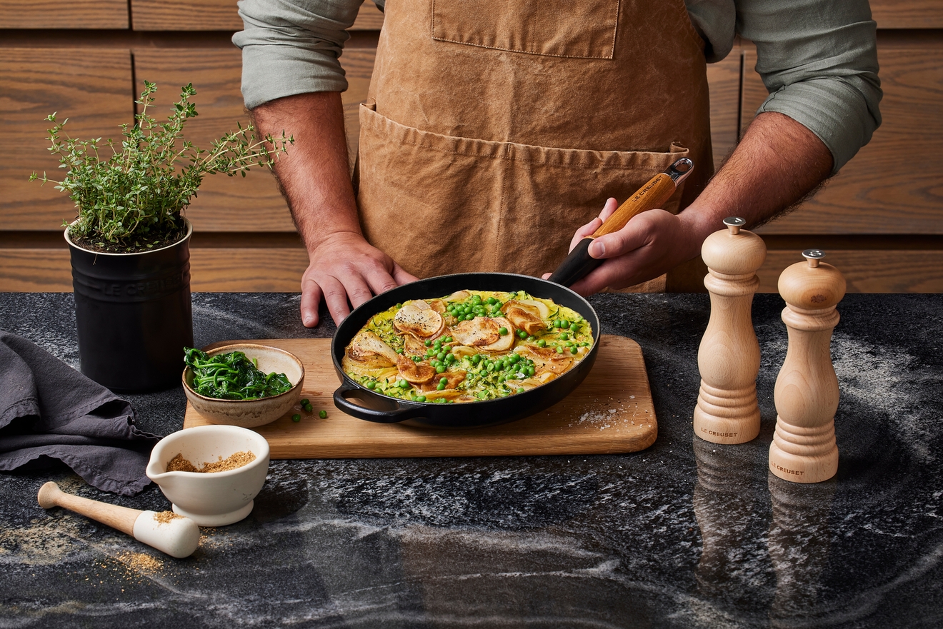 En person i et brunt forkle holder en stekepanne på en kjøkkenbenk omgitt av urte- og krydderkverner.