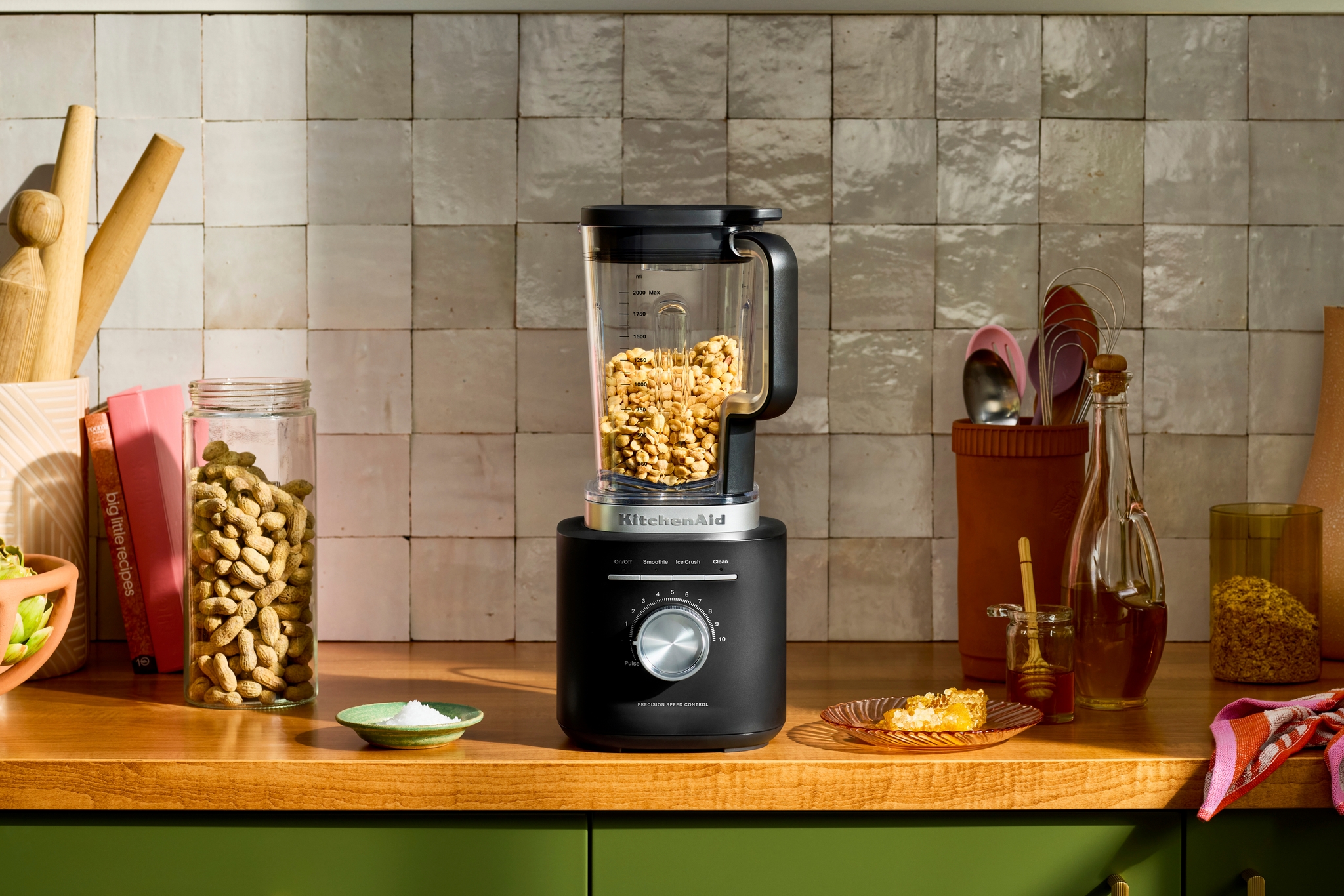 En svart KitchenAid-mixer står på en köksbänk fylld med jordnötter, omgiven av ingredienser och köksredskap.