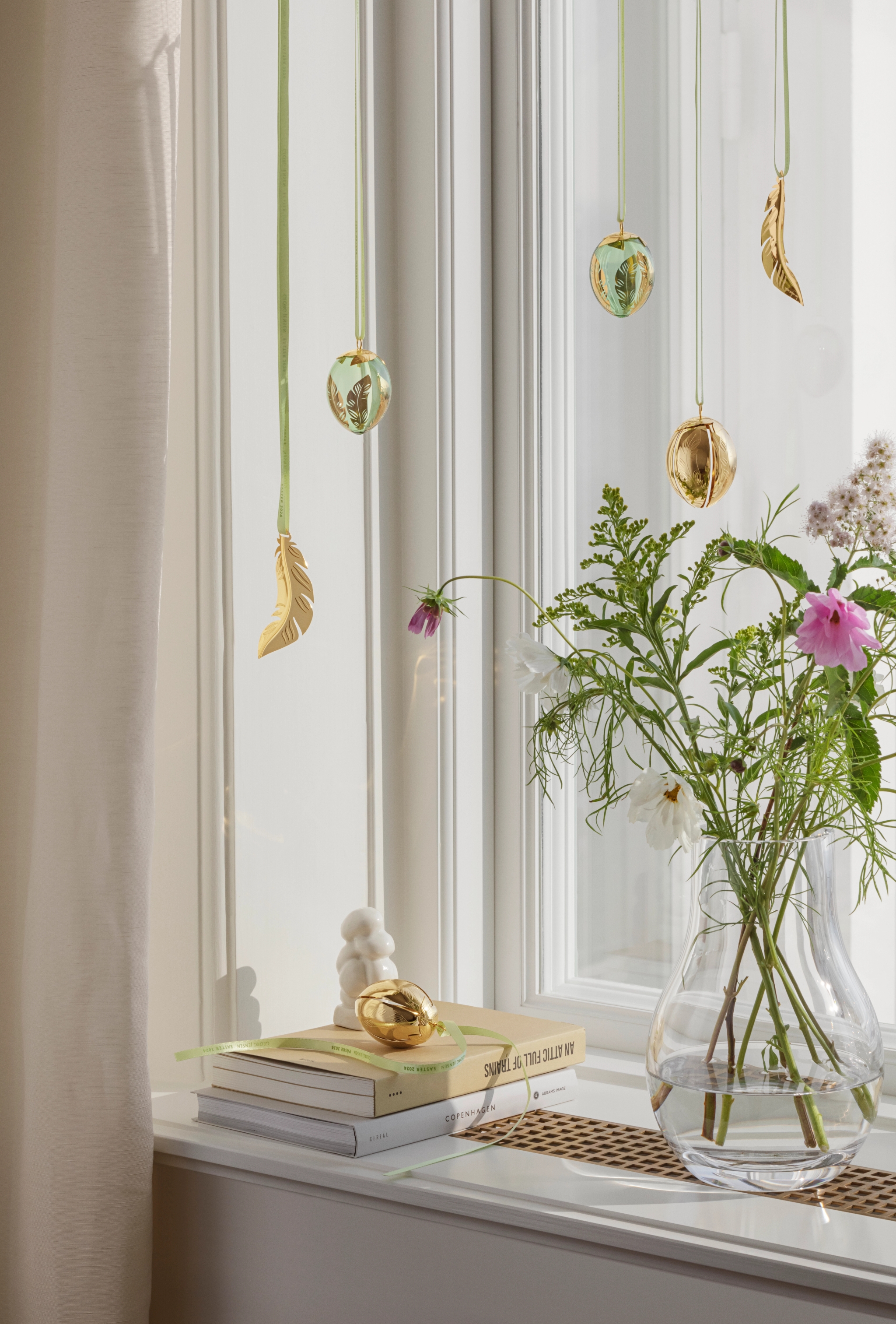 Gullegg- og fjærpynt henger i bånd foran et vindu, med en vase med blomster og bøker i vinduskarmen.