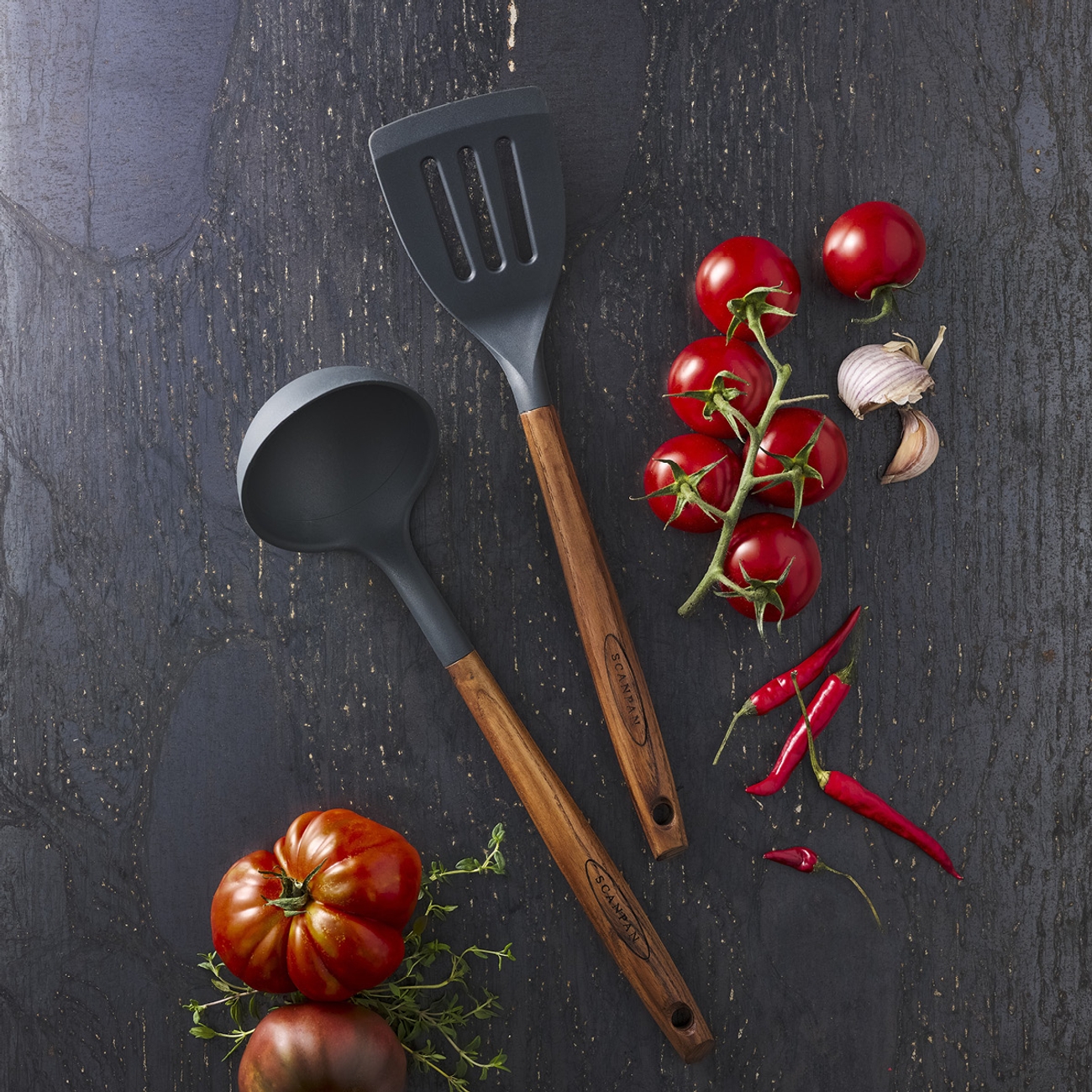 Mörkgrå köksslev och spatel med trähandtag, tillsammans med färska tomater, vitlök och röd chilipeppar på en mörk texturerad yta.