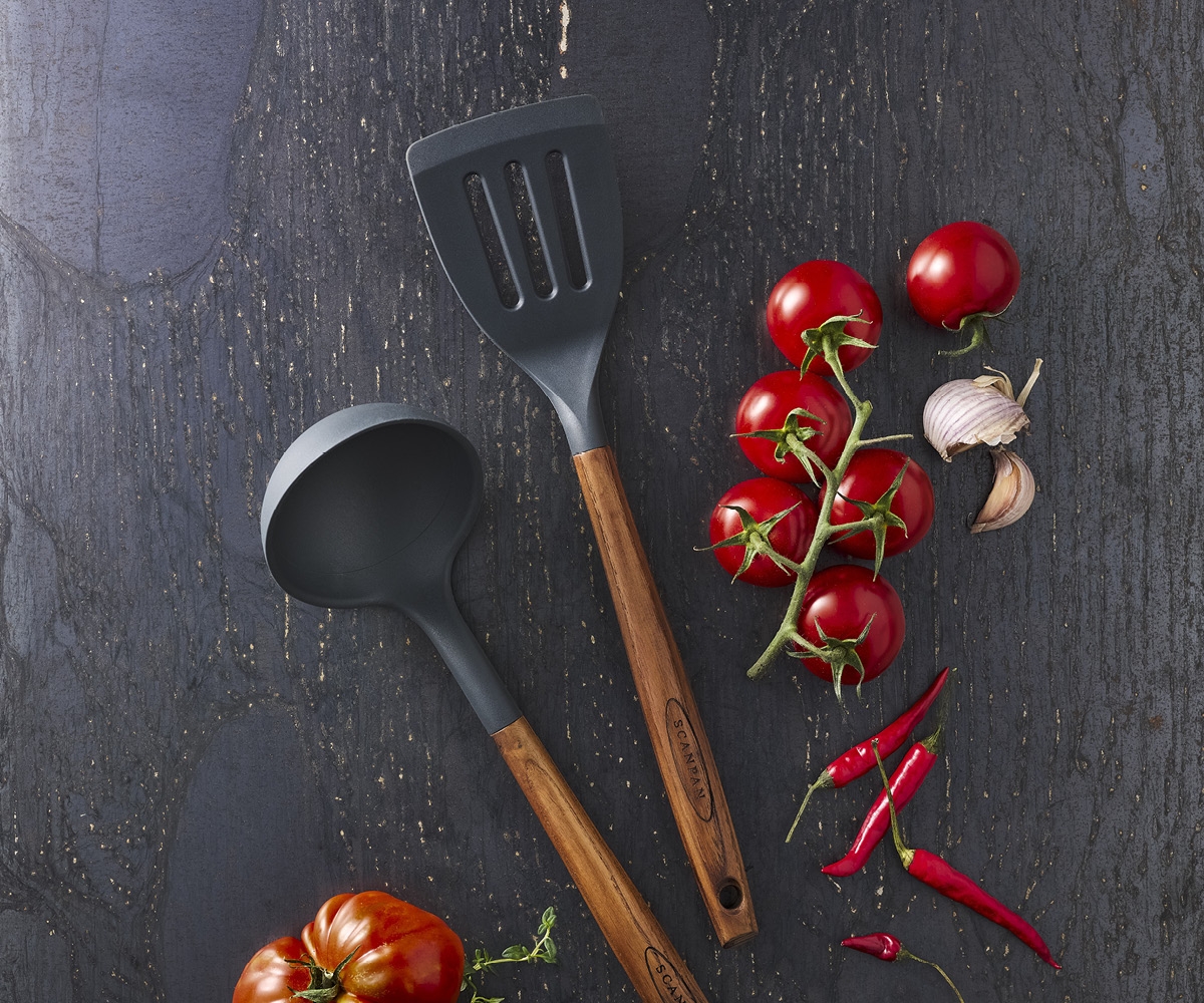 Mörkgrå köksslev och spatel med trähandtag, tillsammans med färska tomater, vitlök och röd chilipeppar på en mörk texturerad yta.