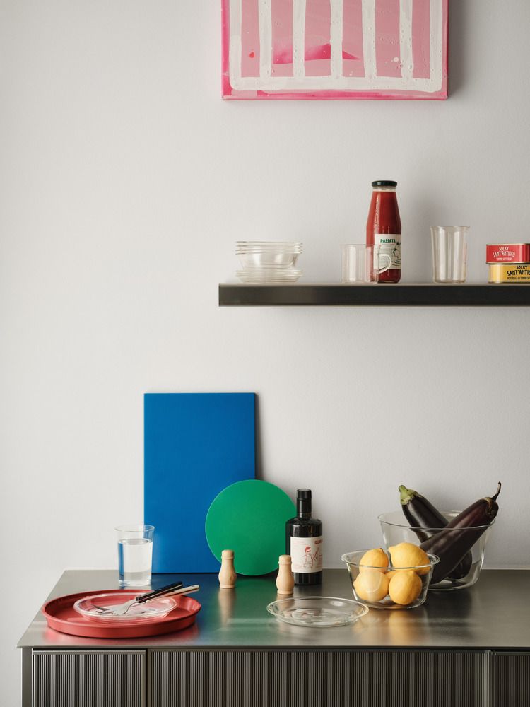 En modern köksscen med en rosa abstrakt målning, en hylla med skafferiartiklar och en bänk med citroner, aubergine, en röd tallrik och färgglada skärbrädor.