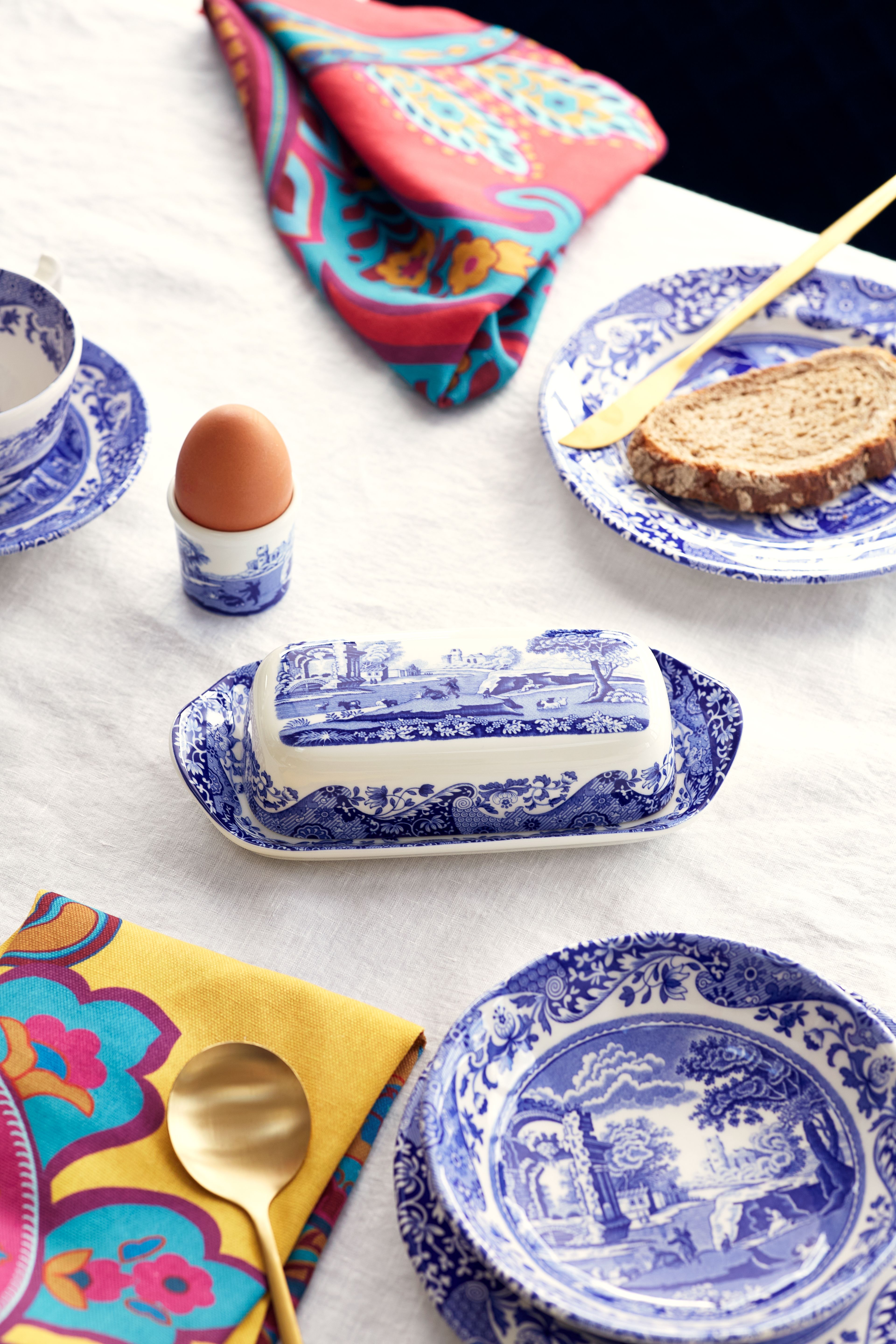 Breakfast table with blue and white patterned dishes, an egg, bread, a butter dish, and colorful napkins on a white tablecloth.