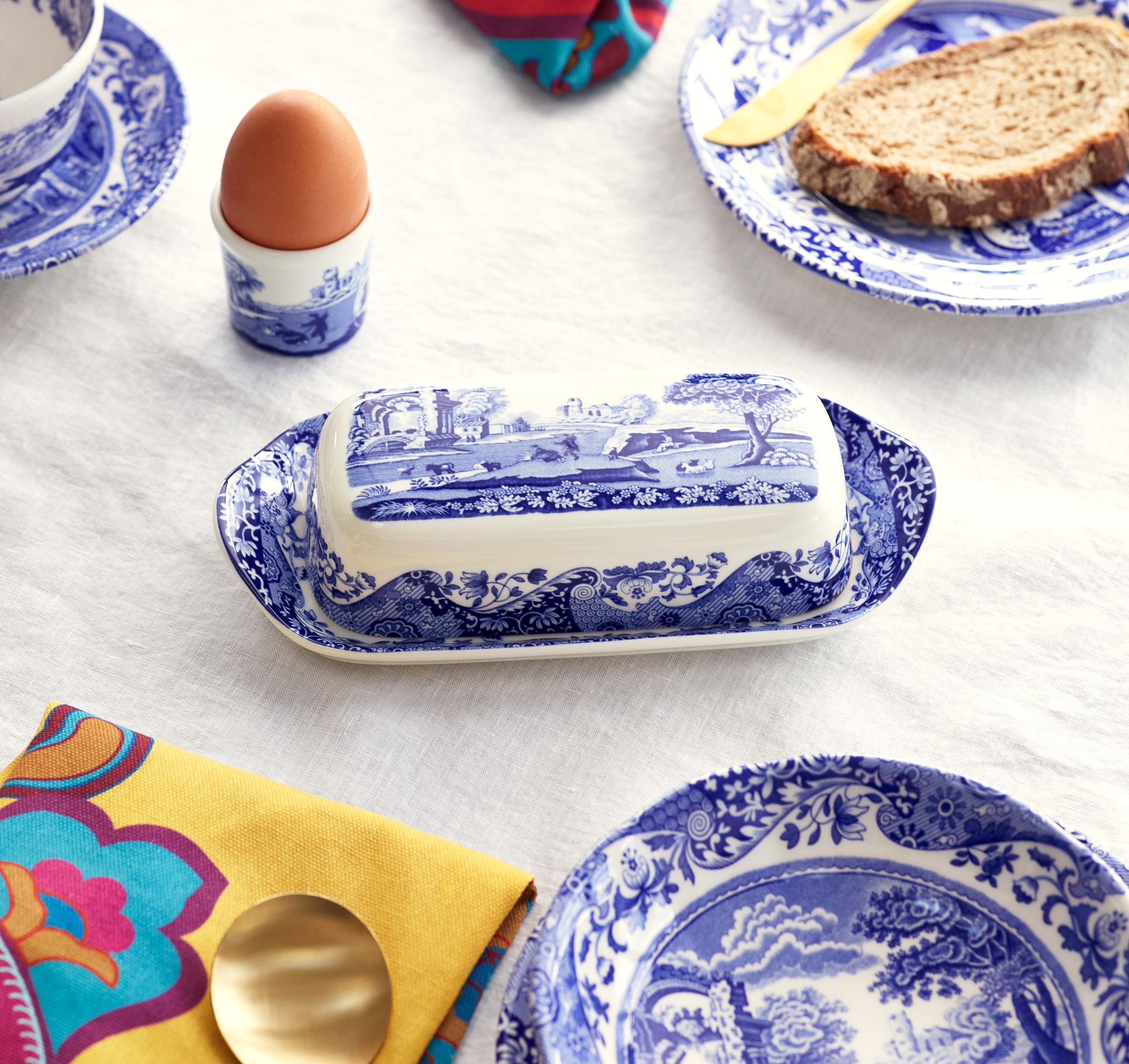 Breakfast table with blue and white patterned dishes, an egg, bread, a butter dish, and colorful napkins on a white tablecloth.
