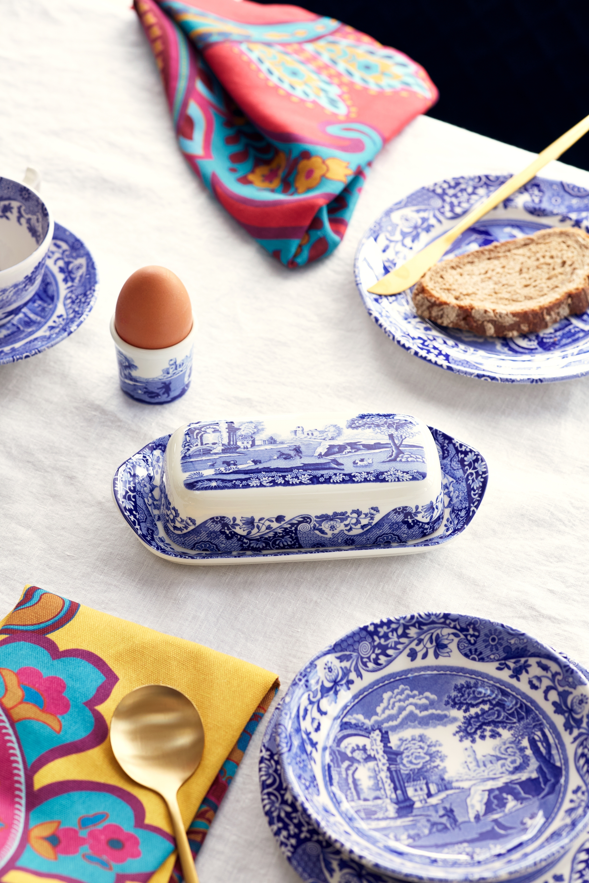 A breakfast spread on a white linen tablecloth, featuring blue and white patterned dishes, a whole egg, a slice of bread, and colorful napkins.