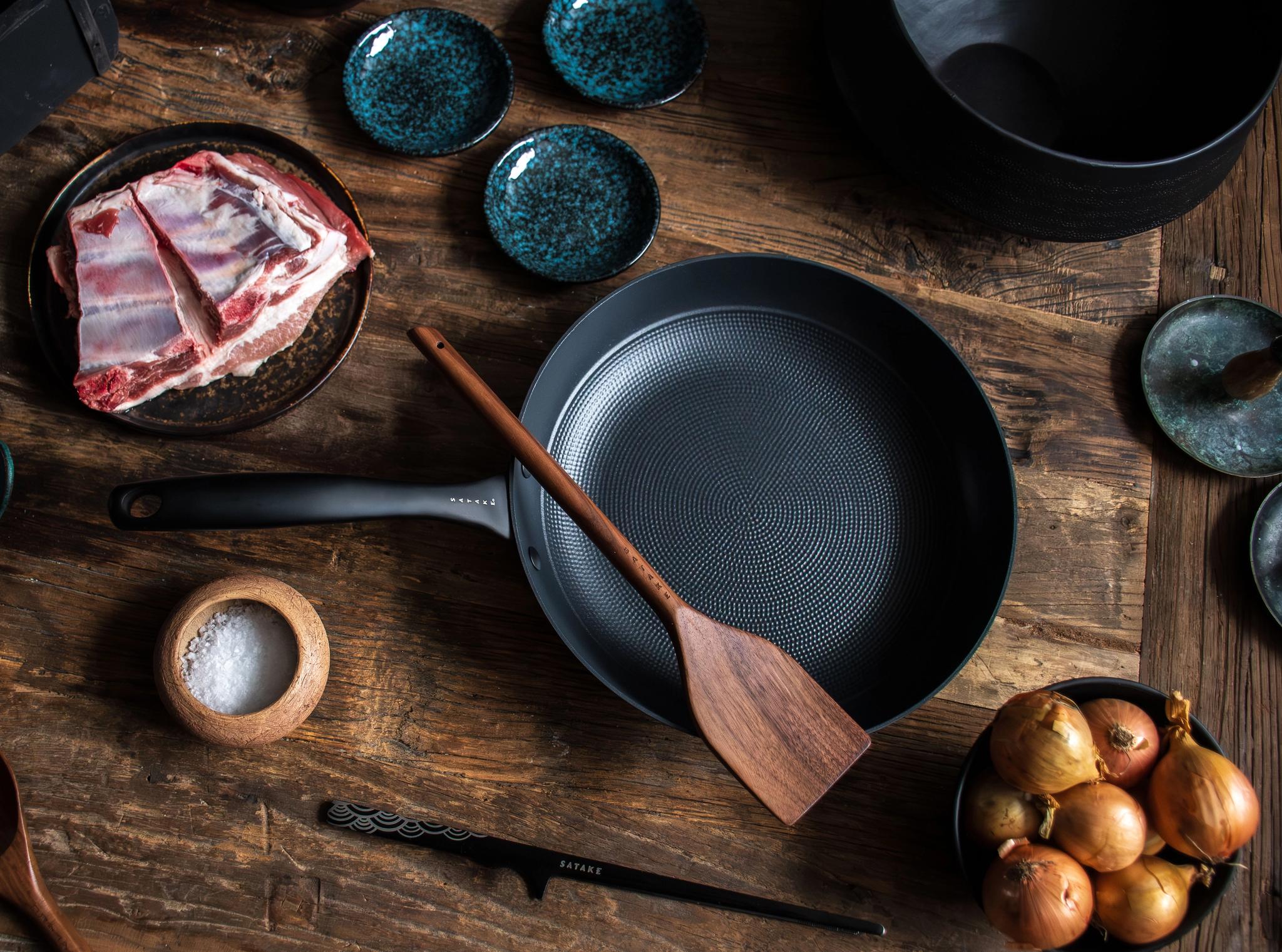Overhead view of raw meat, onions, spices, and a frying pan with a wooden spatula on a rustic wooden table.