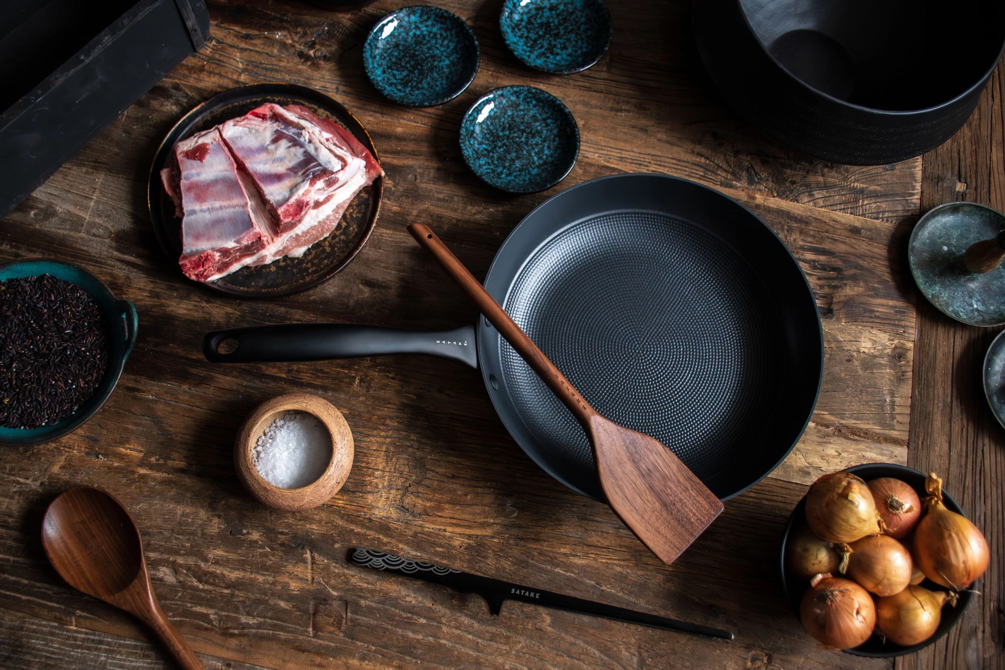 Raw meat, onions, and spices next to a black frying pan and wooden utensils on a rustic wooden table.