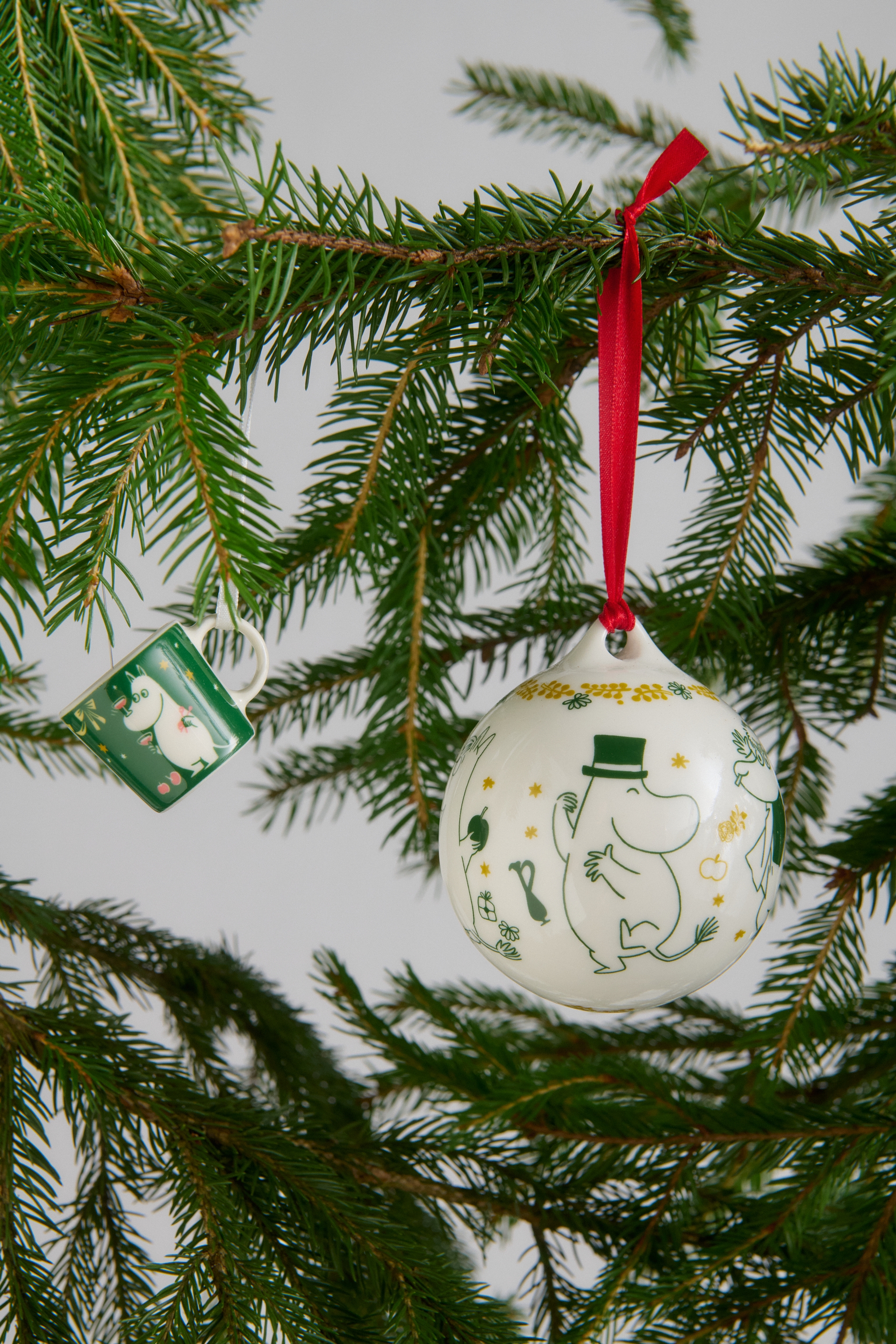 Muminjulgranskula och miniatyrmugg hänger bland gröna grenar i en julgran.