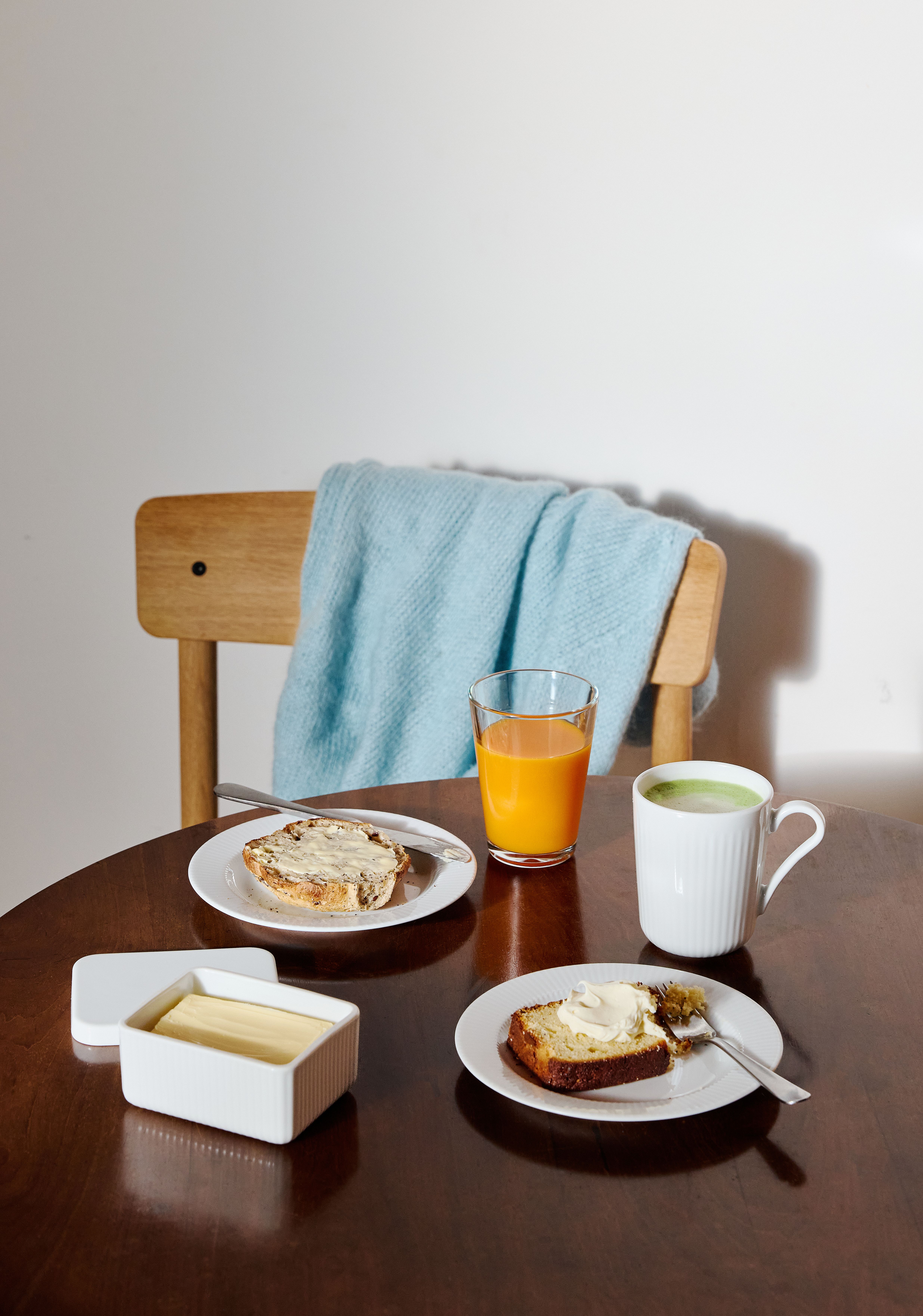 Frukostbord med smörgåsar, apelsinjuice, matcha och smör framför en trästol med en ljusblå filt.