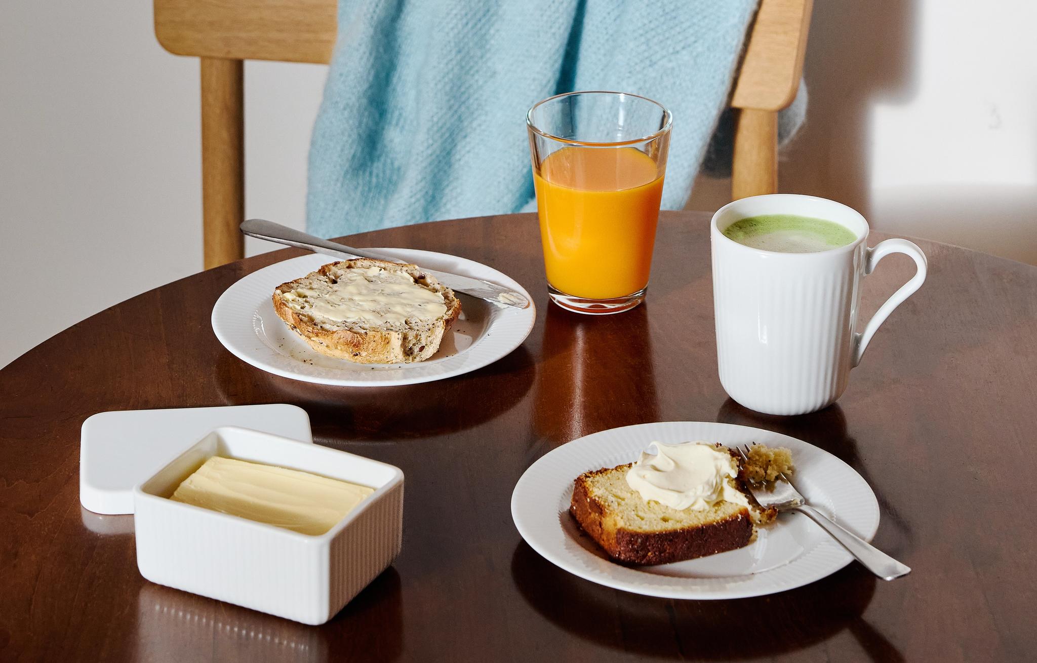 Frukostbord med smörgåsar, apelsinjuice, matcha och smör framför en trästol med en ljusblå filt.