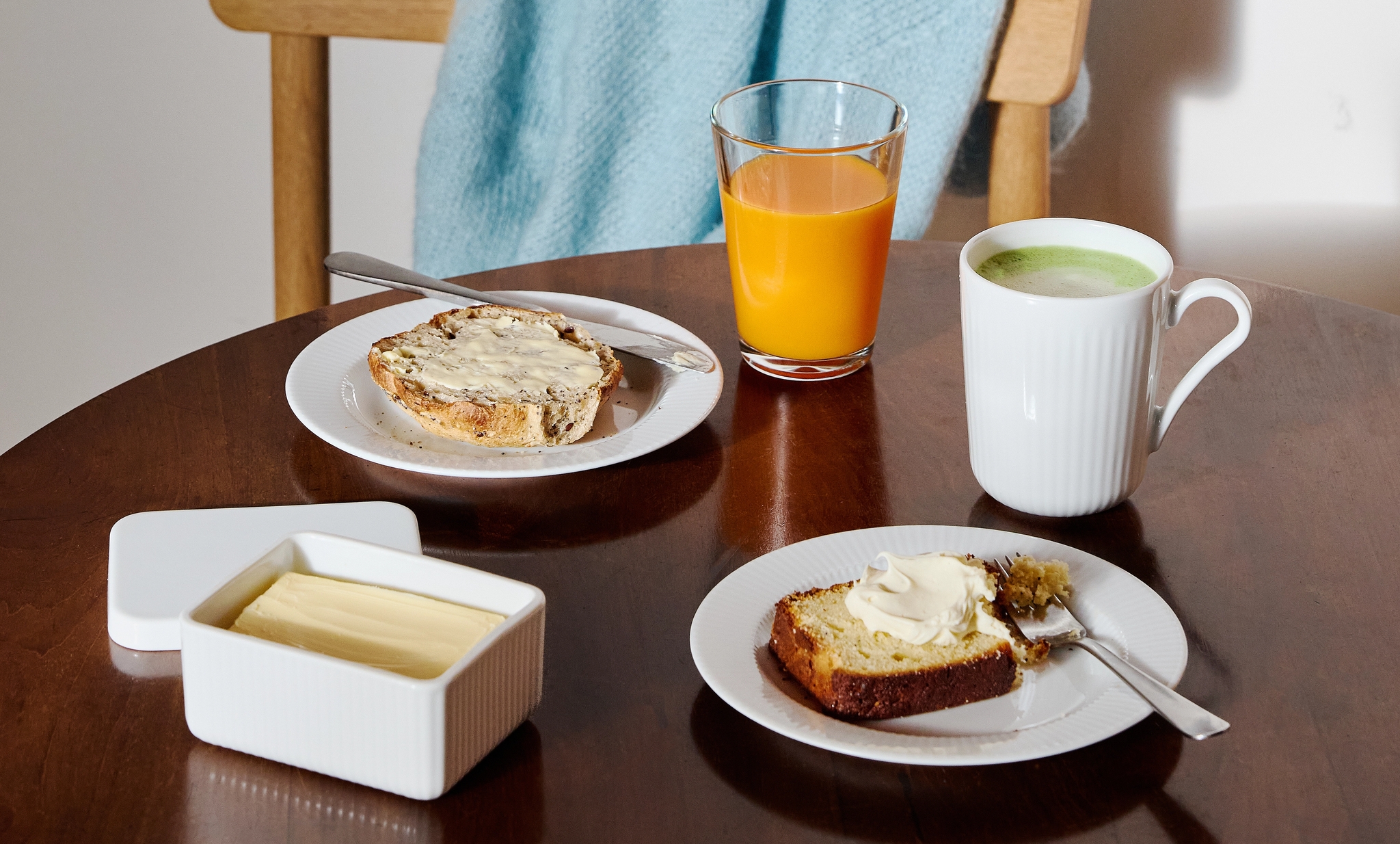 Frukostbord med smörgåsar, apelsinjuice, matcha och smör framför en trästol med en ljusblå filt.