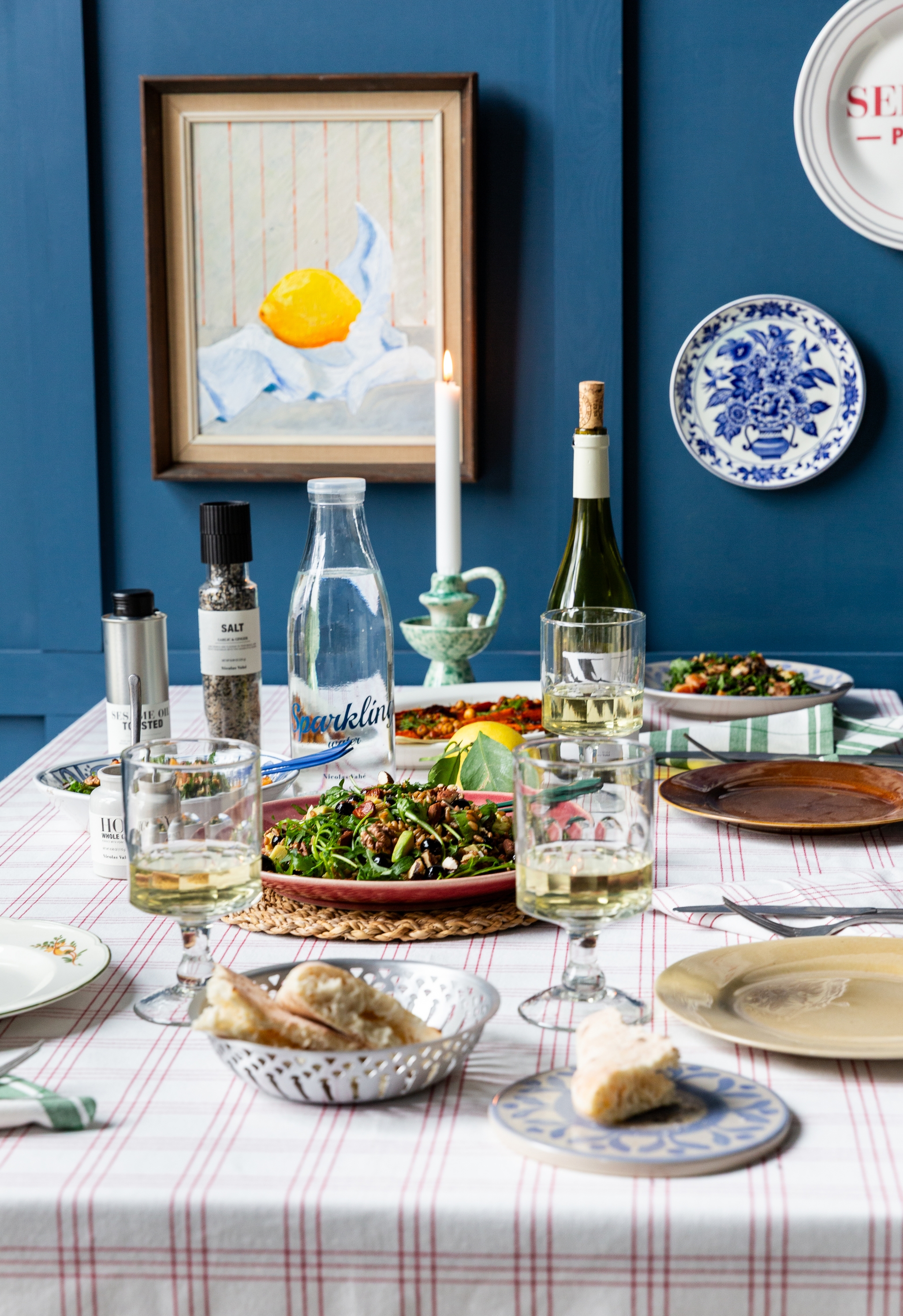 Ett matbord dukat med sallader, vitt vin och bröd, mot en blå vägg dekorerad med en citronmålning och dekorativa tallrikar