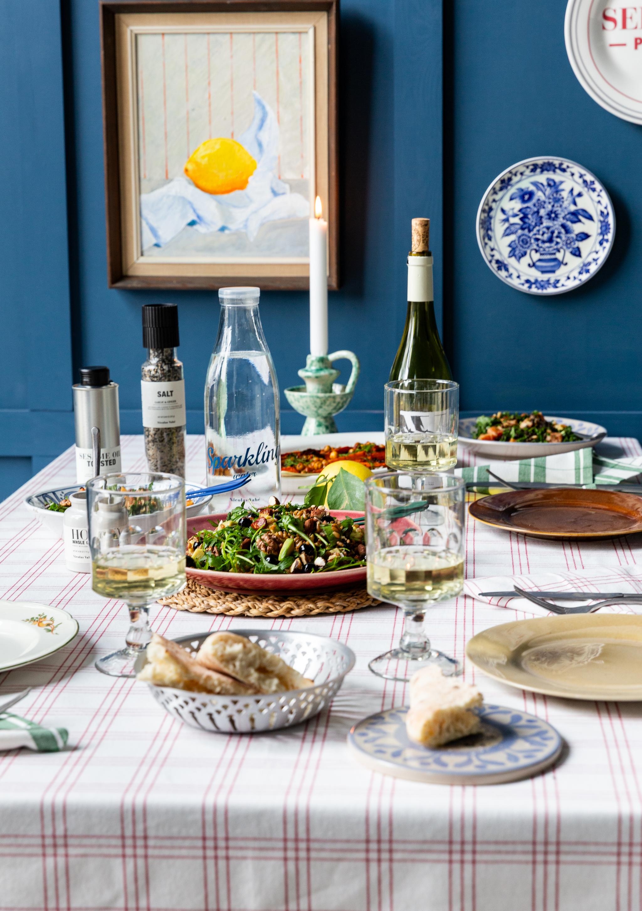 Ett matbord dukat med sallader, vitt vin och bröd, mot en blå vägg dekorerad med en citronmålning och dekorativa tallrikar