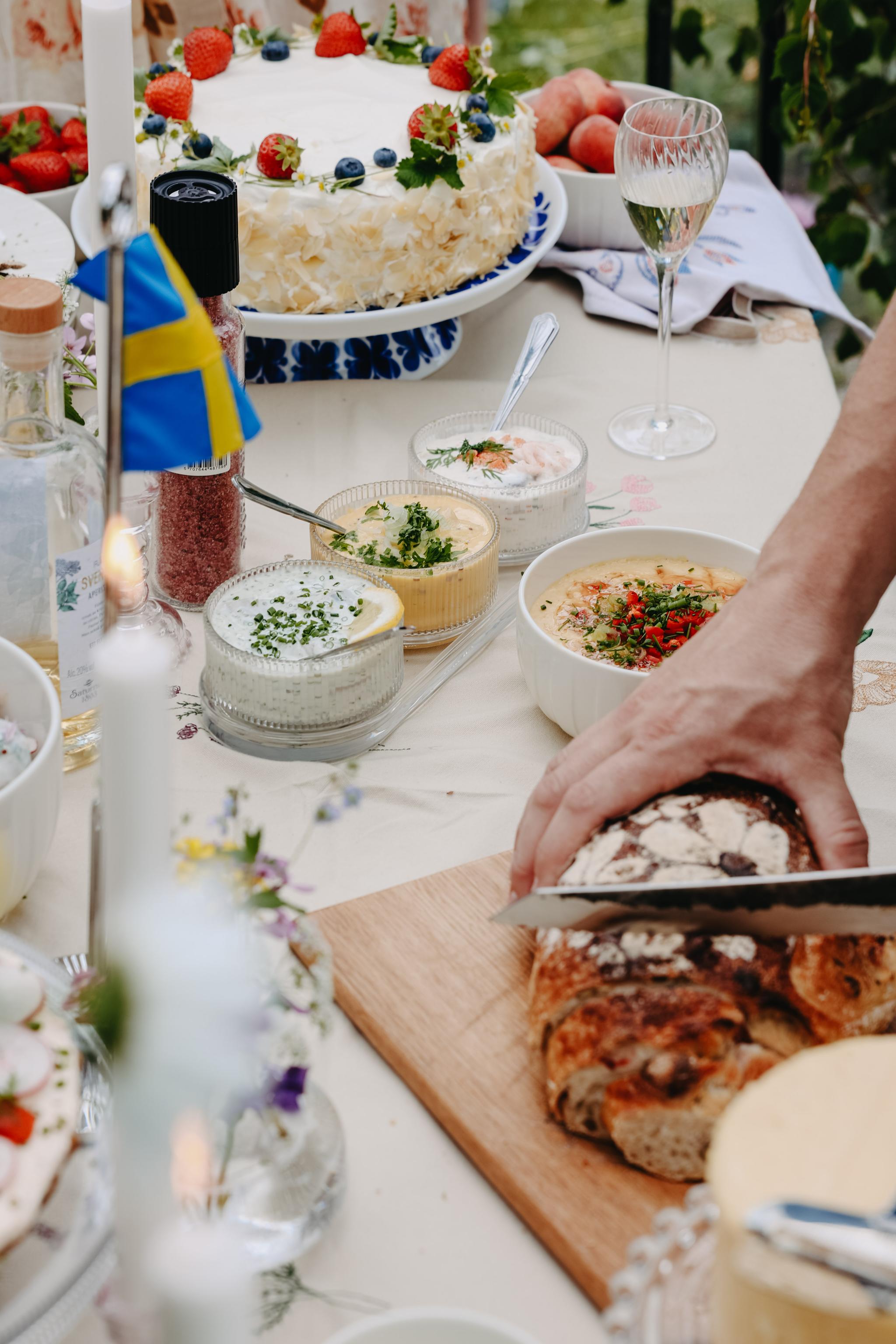 Somrig svensk festdukning med tårta, sillröror och nybakat bröd som skärs upp på ett bord dekorerat med flagga och blommor.