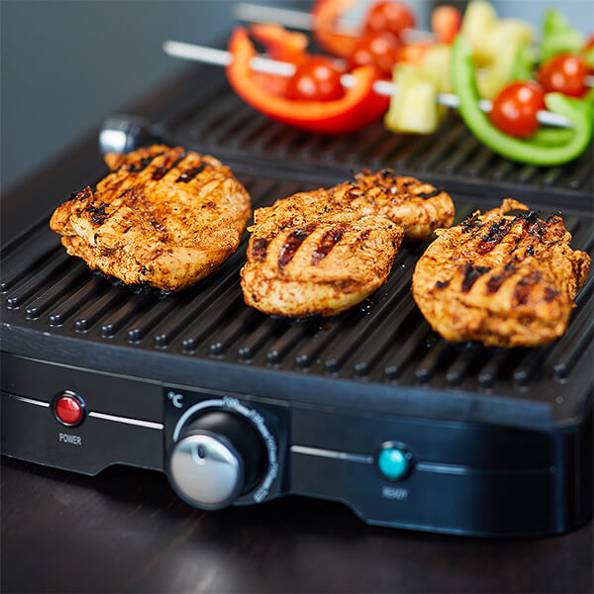 Sähkögrillillä paistuu mehukkaita kananrintafileitä ja taustalla näkyy värikkäitä kasvisvartaita.