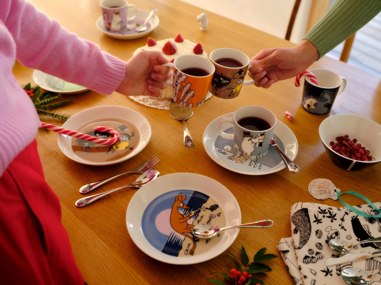 Två personer skålar i Muminmuggar vid ett träbord dukat med festliga Mumin-fat, en kaka och polkagrisar.