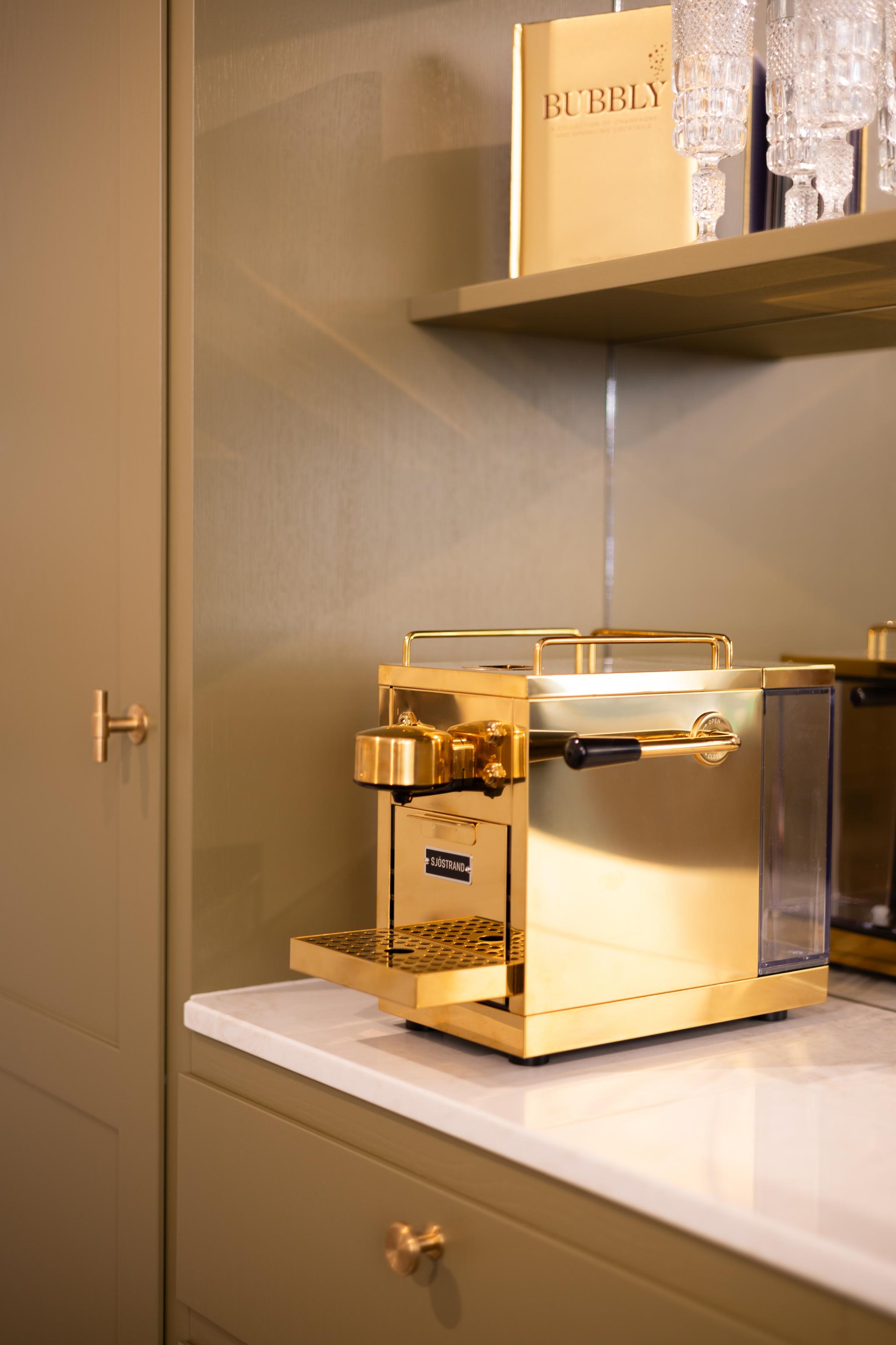 En guldfärgad espressomaskin står på en vit bänkskiva, omgiven av olivgröna skåp med guldfärgade beslag. En "Bubbly"-bok och glas står på en hylla ovanför.