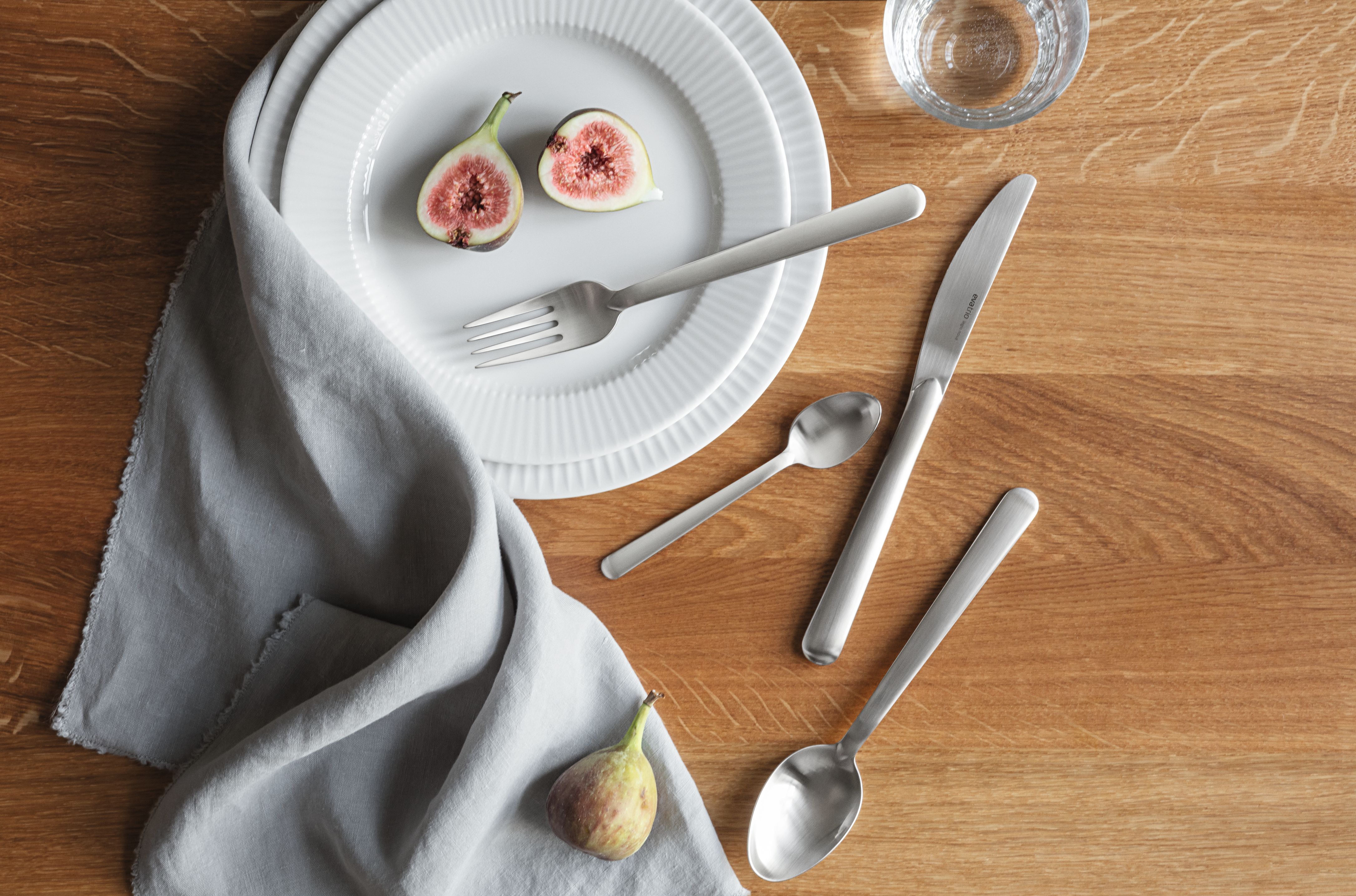  Bild ovanifrån av en dukning med en vit tallrik, skivade fikon, en grå servett och silverbestick på ett träbord