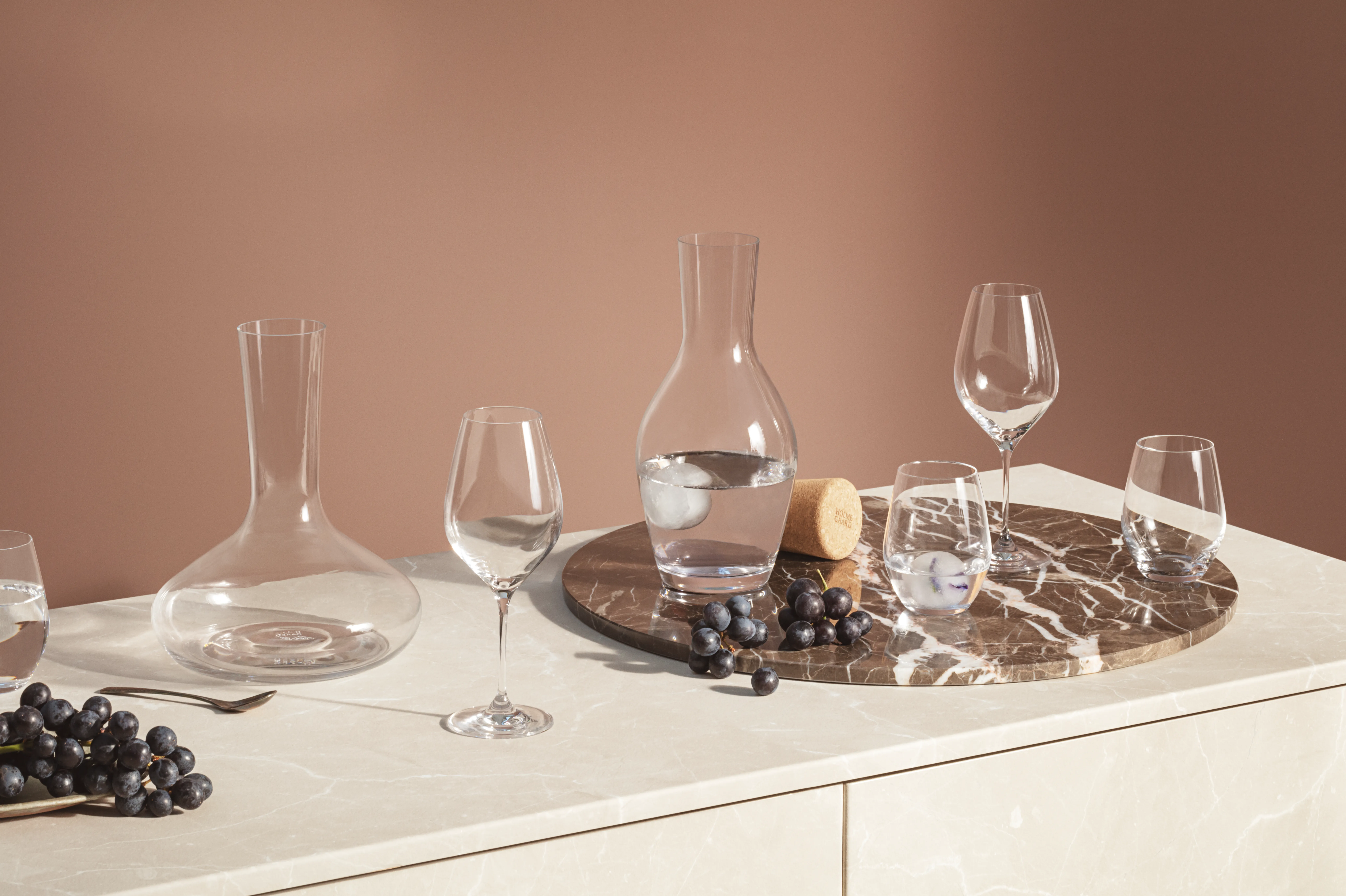 Ulike klare glasskarafler, vinglass og drikkeglass, noen med vann og is, på en marmordisk med mørke druer