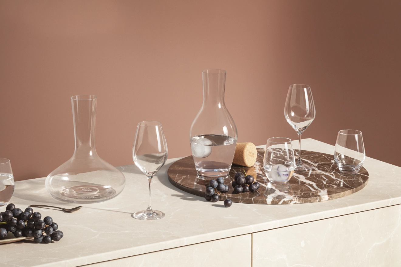 Ulike klare glasskarafler, vinglass og drikkeglass, noen med vann og is, på en marmordisk med mørke druer
