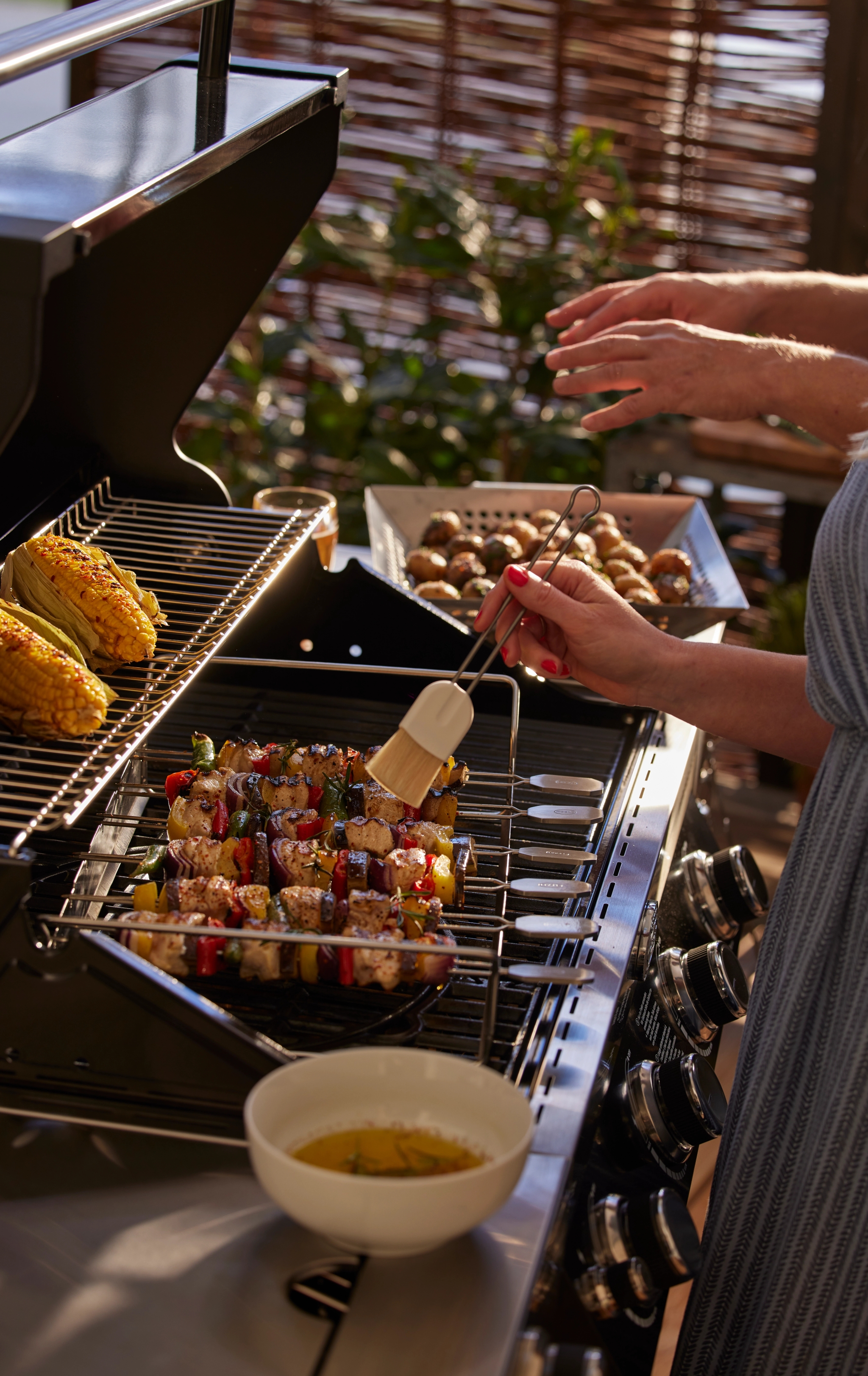 En kvinna penslar kyckling- och grönsaksspett på en gasolgrill, medan majskolvar tillagas bredvid.