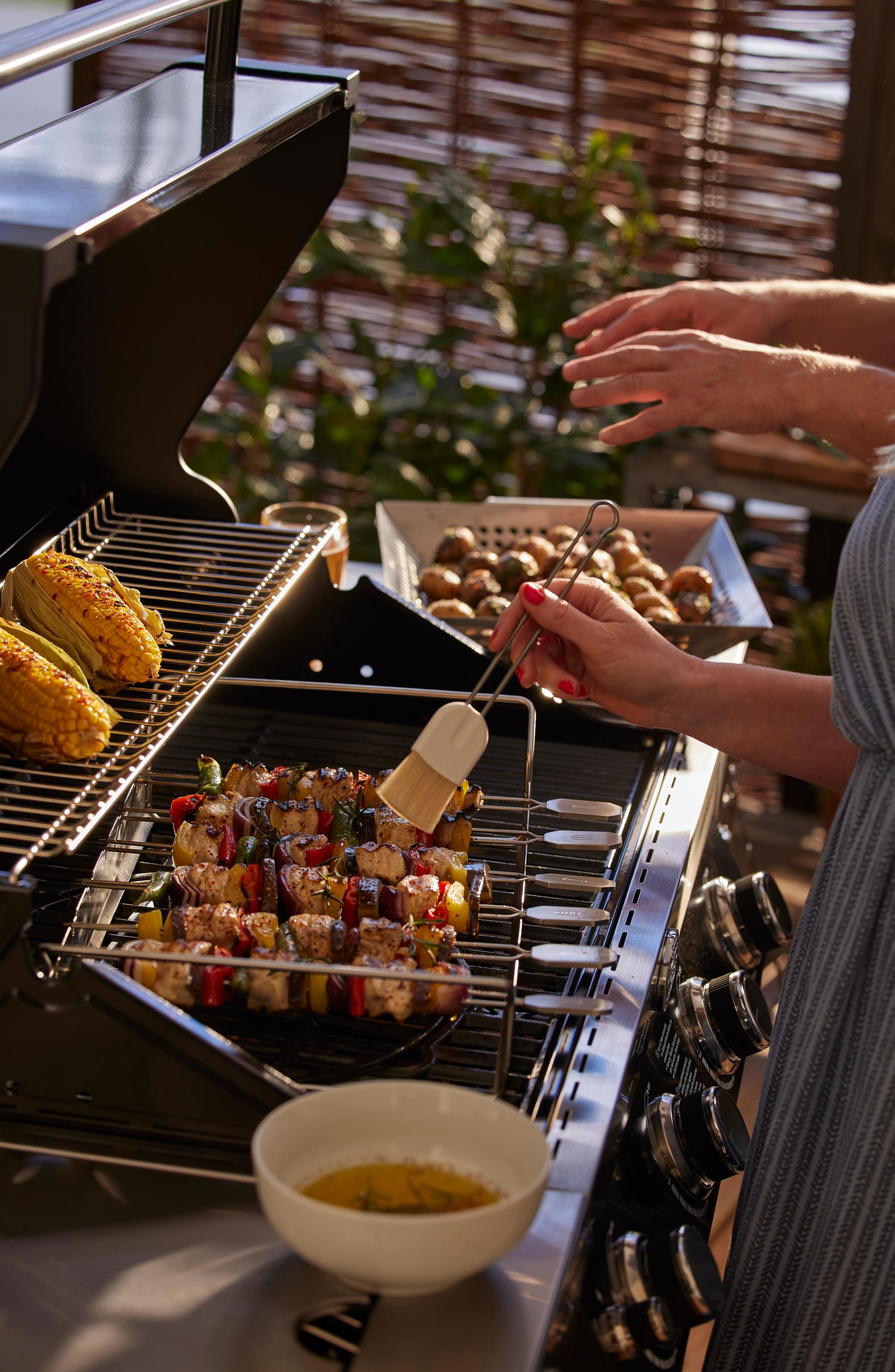 En person penslar färgglada kyckling- och grönsaksspett på en grill, med majskolvar och ugnsrostad potatis