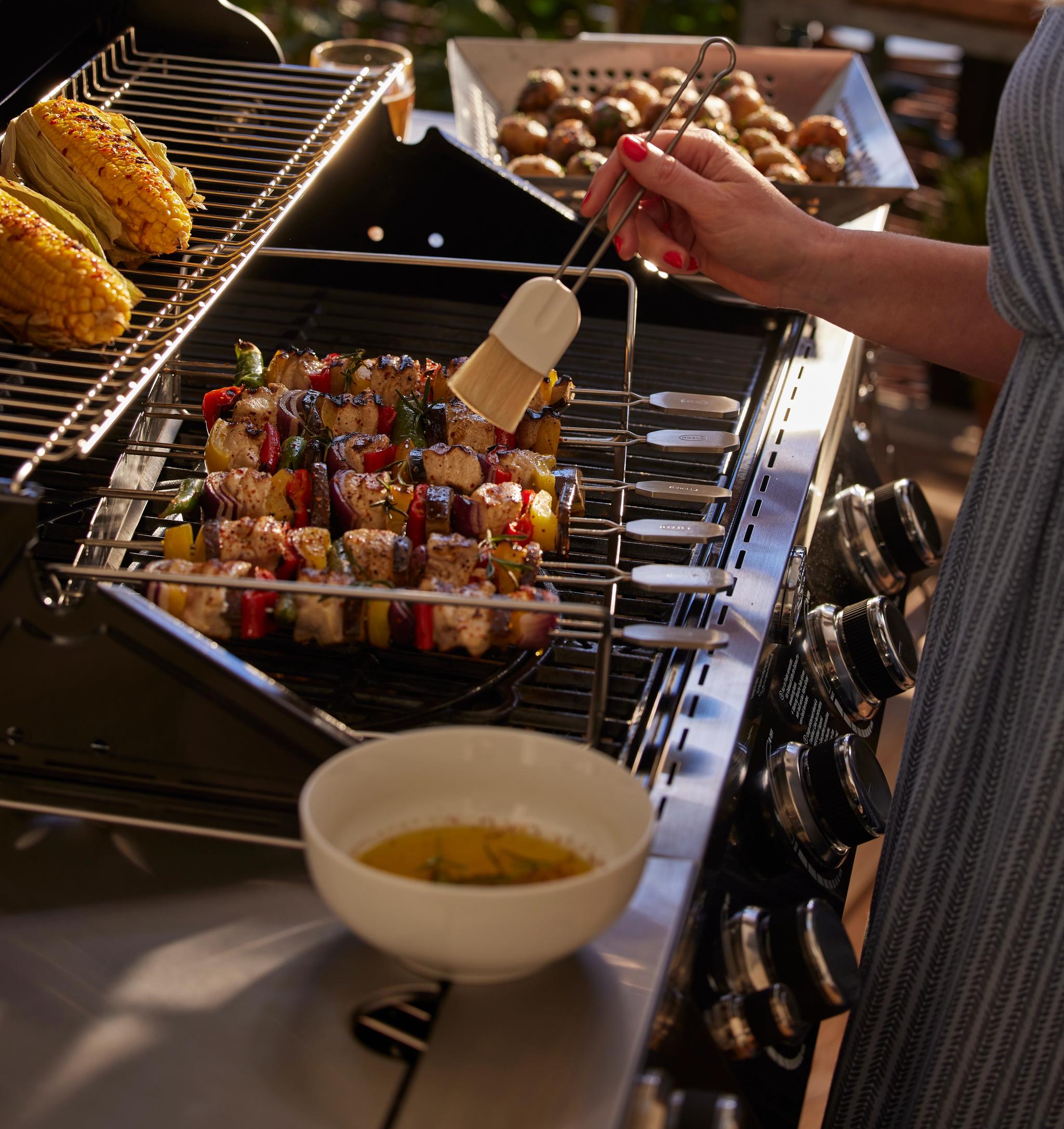 En person penslar kyckling- och grönsaksspett på en grill, med majskolvar och en skål med sås i närheten