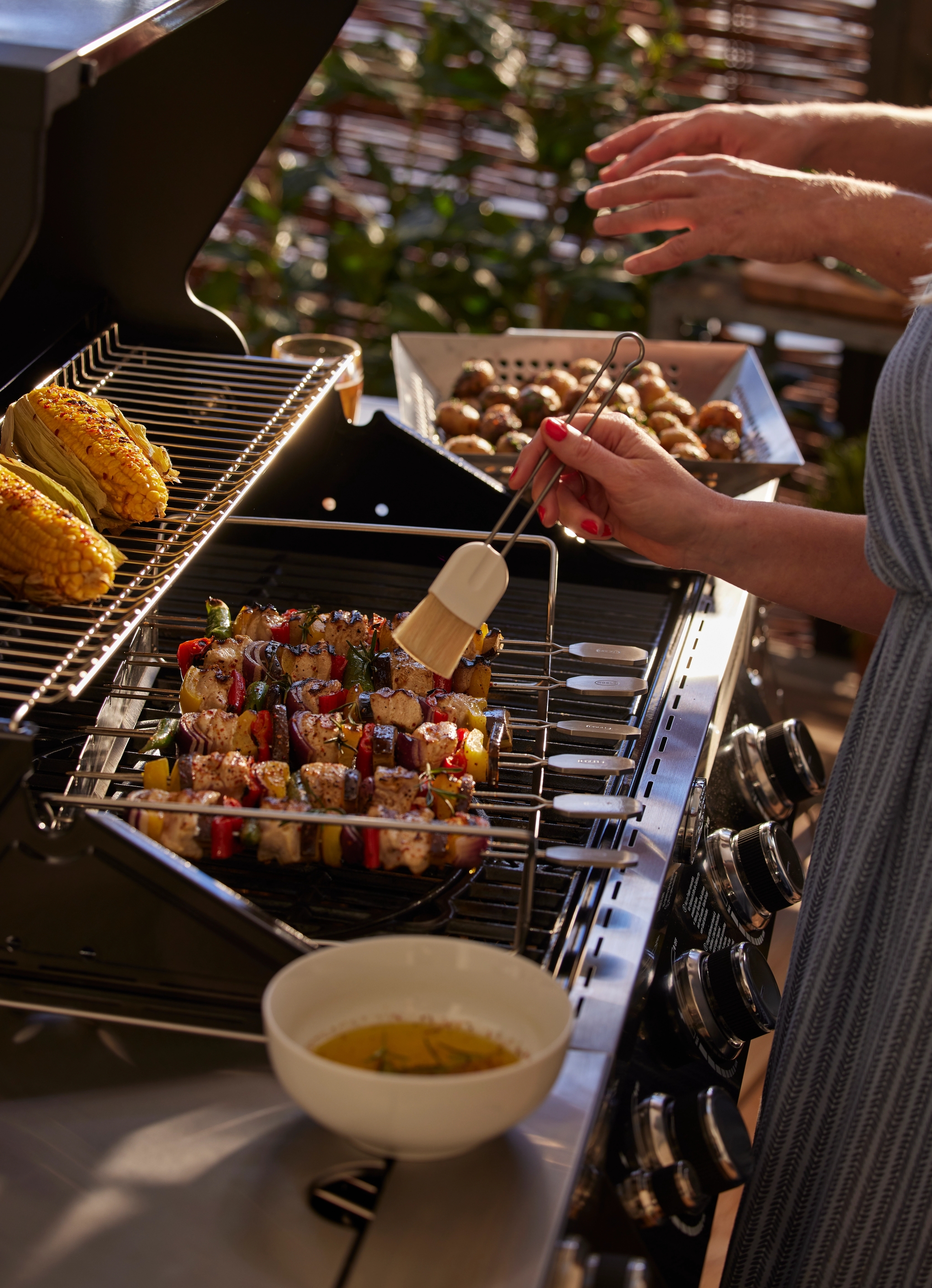 En person penslar kyckling- och grönsaksspett på en grill, med majskolvar och en skål med sås i närheten