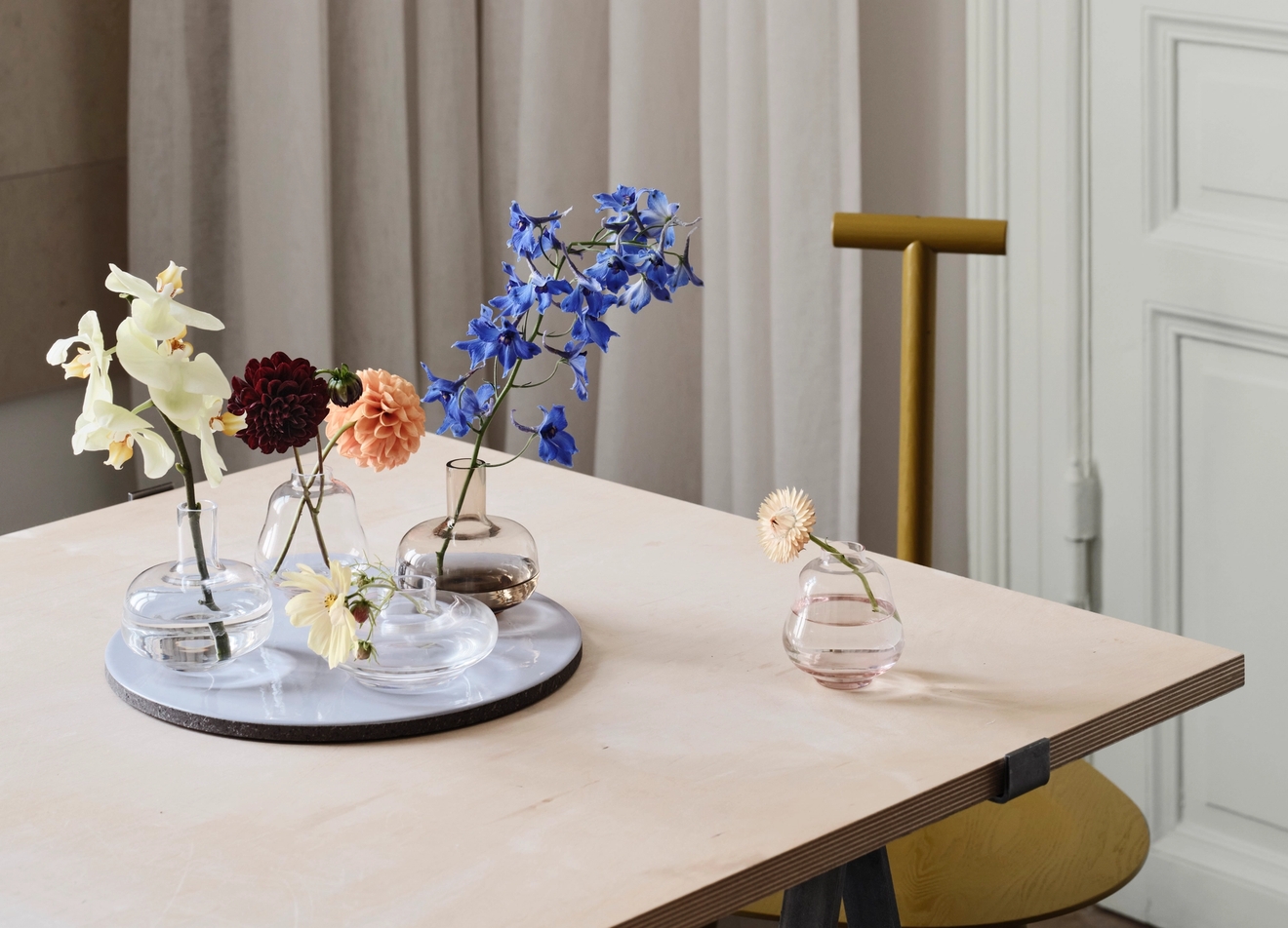 Ulike blomster i glassvaser og på et hvitt brett, plassert på et trebord.