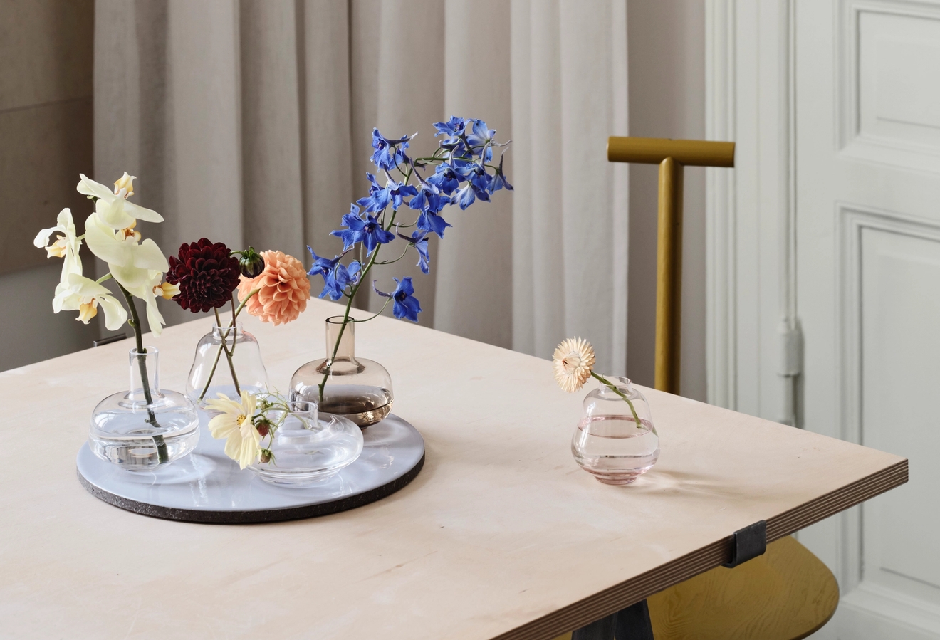 Olika blommor i glasvaser och på en vit bricka, placerade på ett träbord.