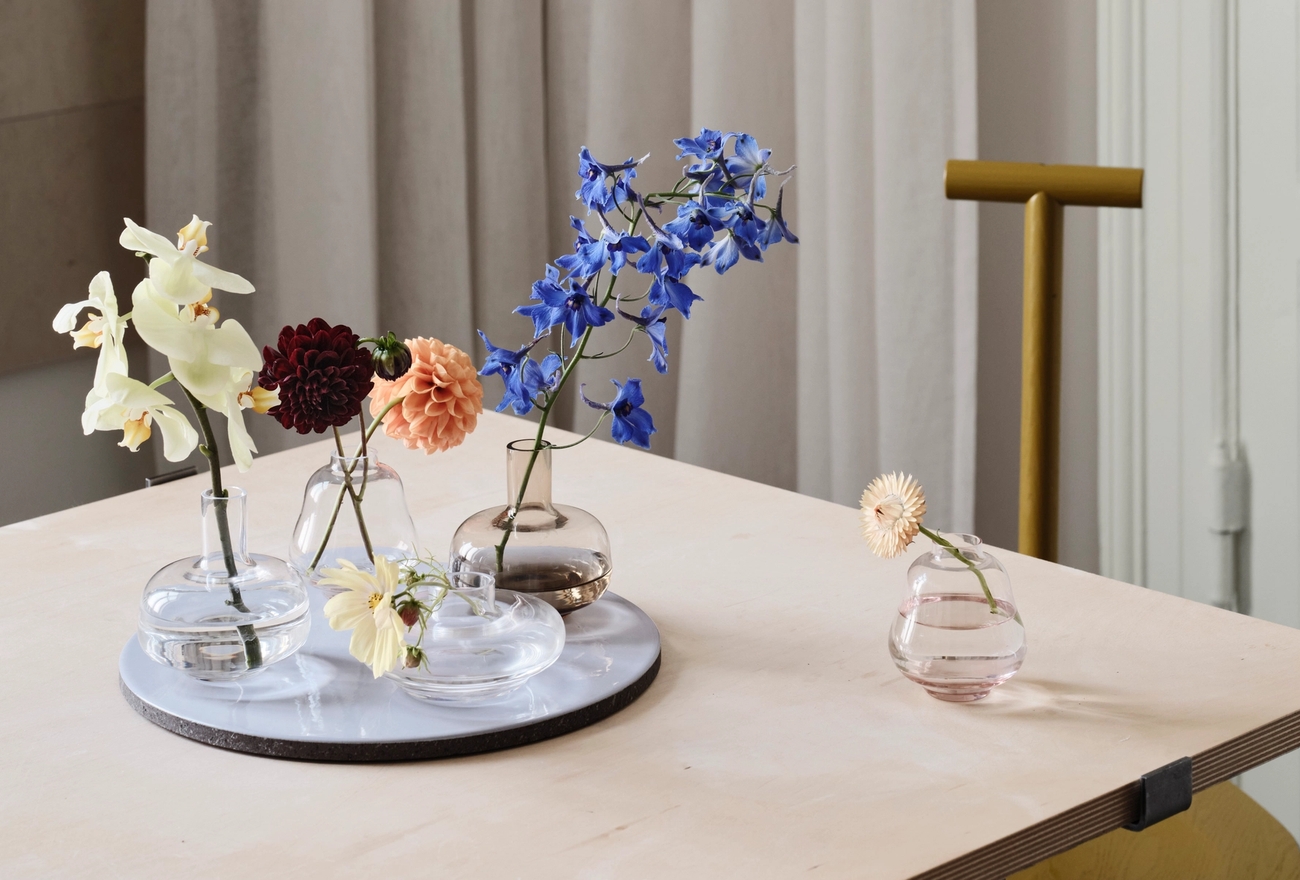 Olika blommor i glasvaser och på en vit bricka, placerade på ett träbord.