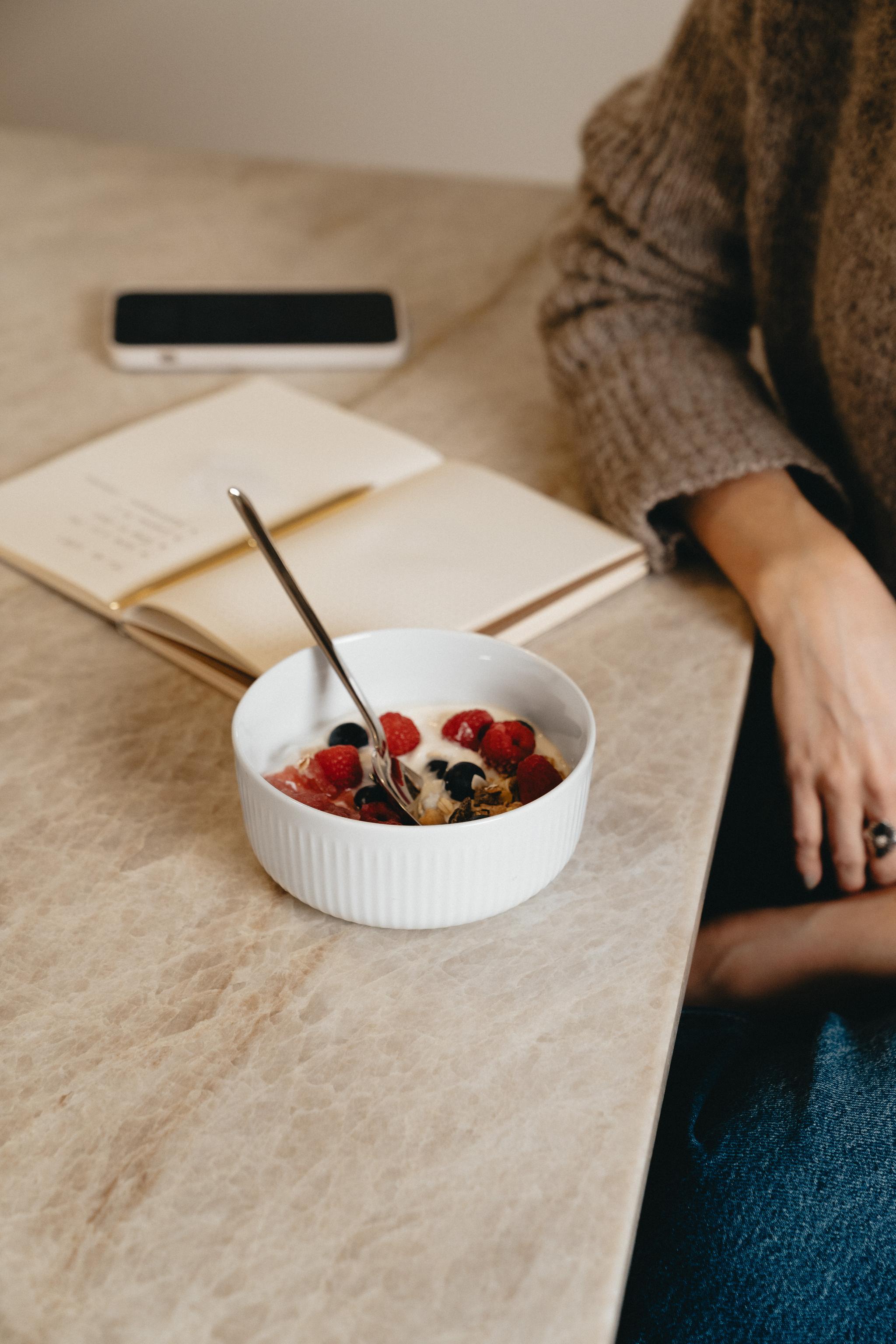 Pöydällä kulhollinen jogurttia marjojen ja lusikan kanssa, vieressä henkilö, avoin muistikirja ja puhelin.