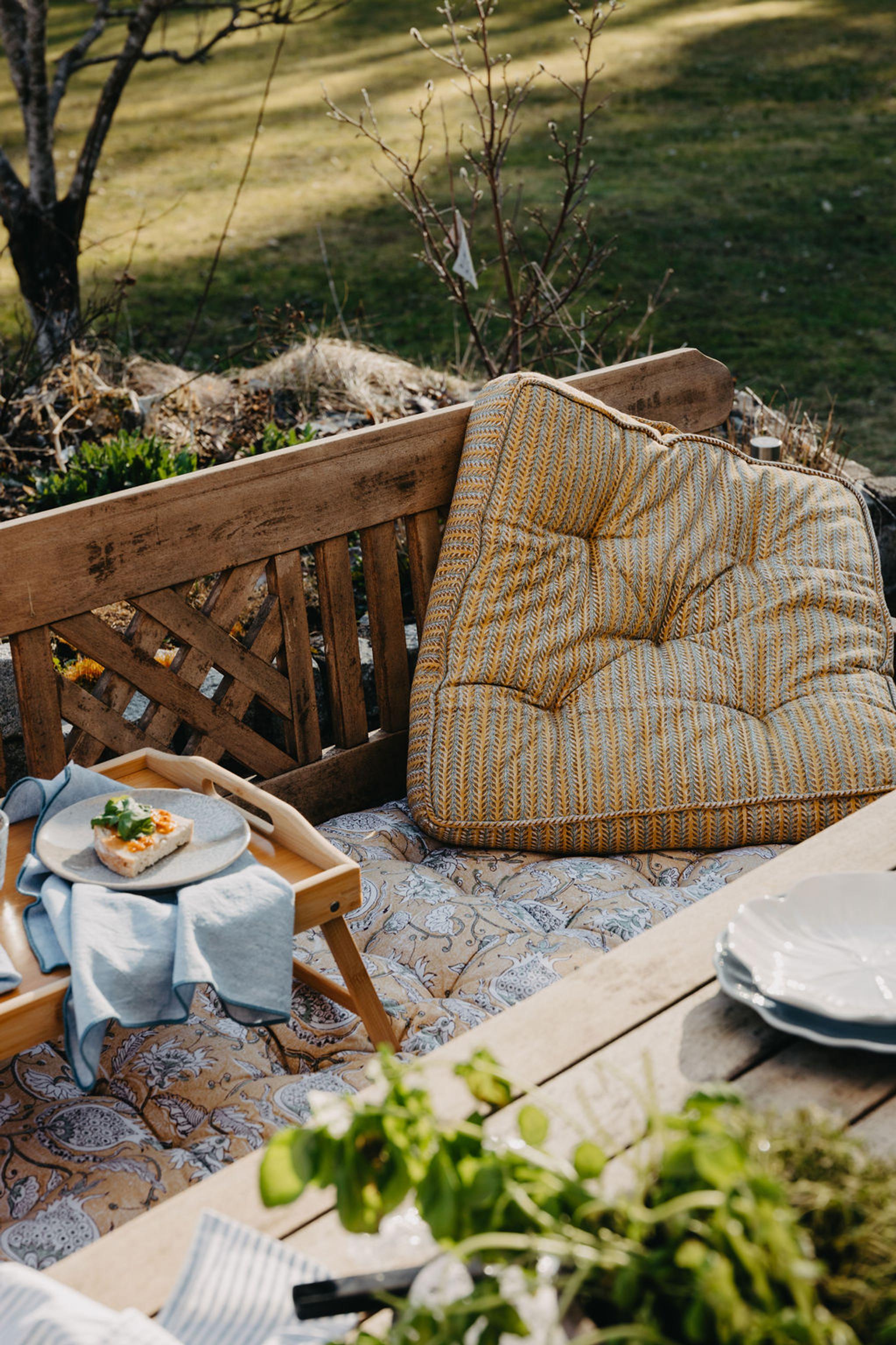 Mysig utomhusmiljö med en träbänk, mönstrade kuddar, en bricka med en öppen smörgås och ett bord med tallrikar och örter.