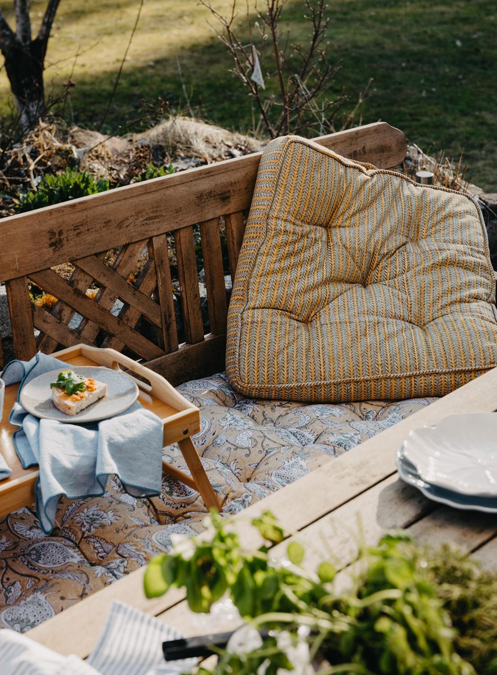 Koselig uteplass med en trebenk, mønstrede puter, et frokostbrett med en liten matbit og friske urter på et bord