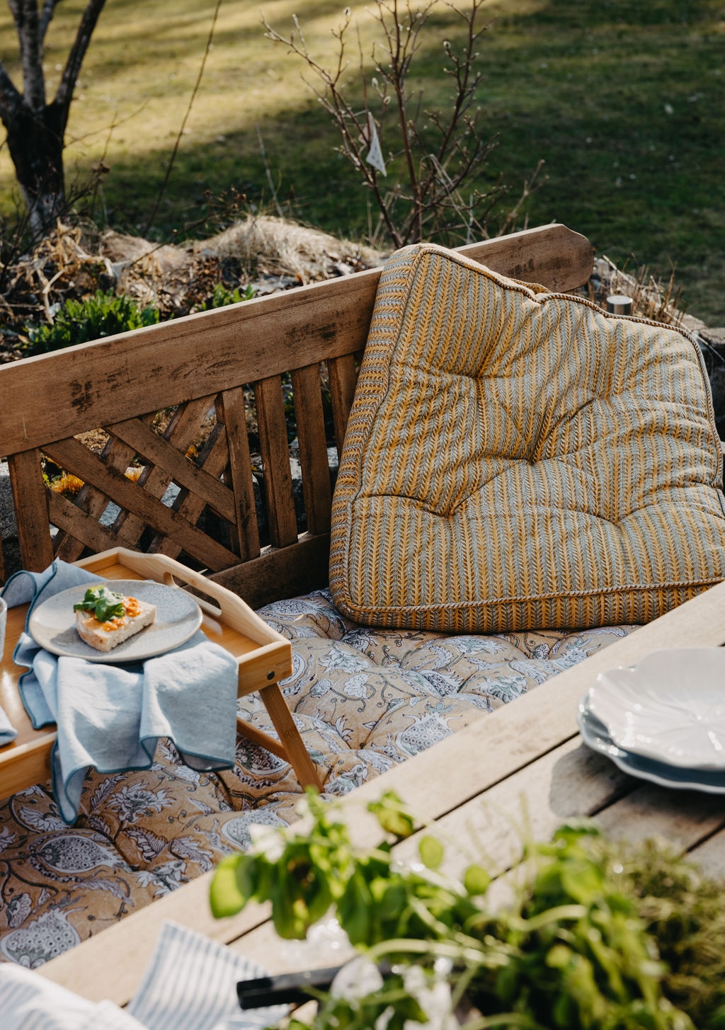 Koselig uteplass med en trebenk, mønstrede puter, et frokostbrett med en liten matbit og friske urter på et bord