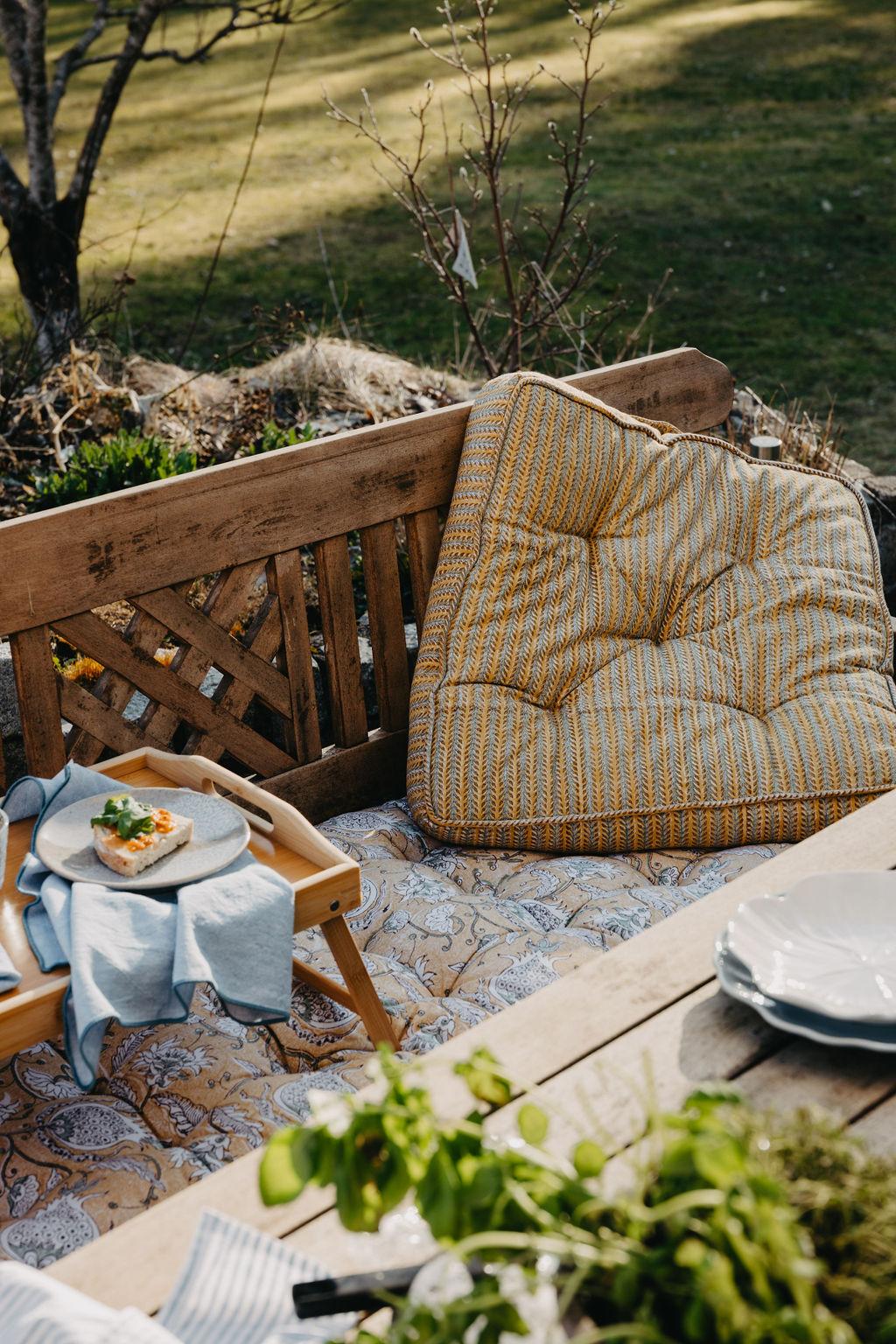 Mysig utomhusmiljö med en träbänk, mönstrade kuddar, en bricka med en öppen smörgås och ett bord med tallrikar och örter.
