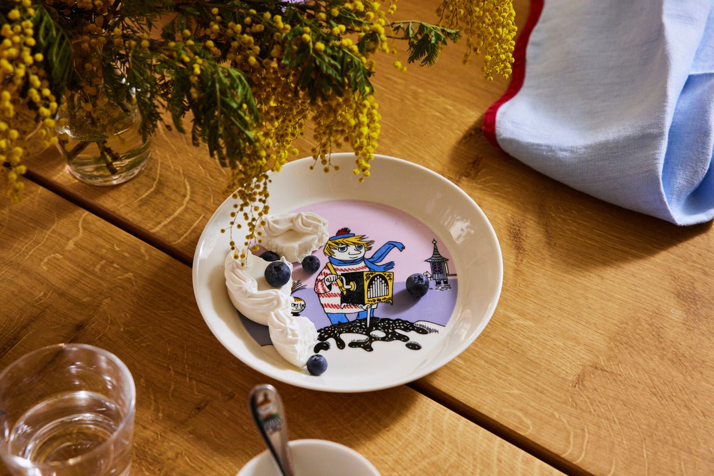 En barnsligt illustrerad tallrik med vispad grädde och blåbär står på ett dukat träbord bredvid gula blommor.