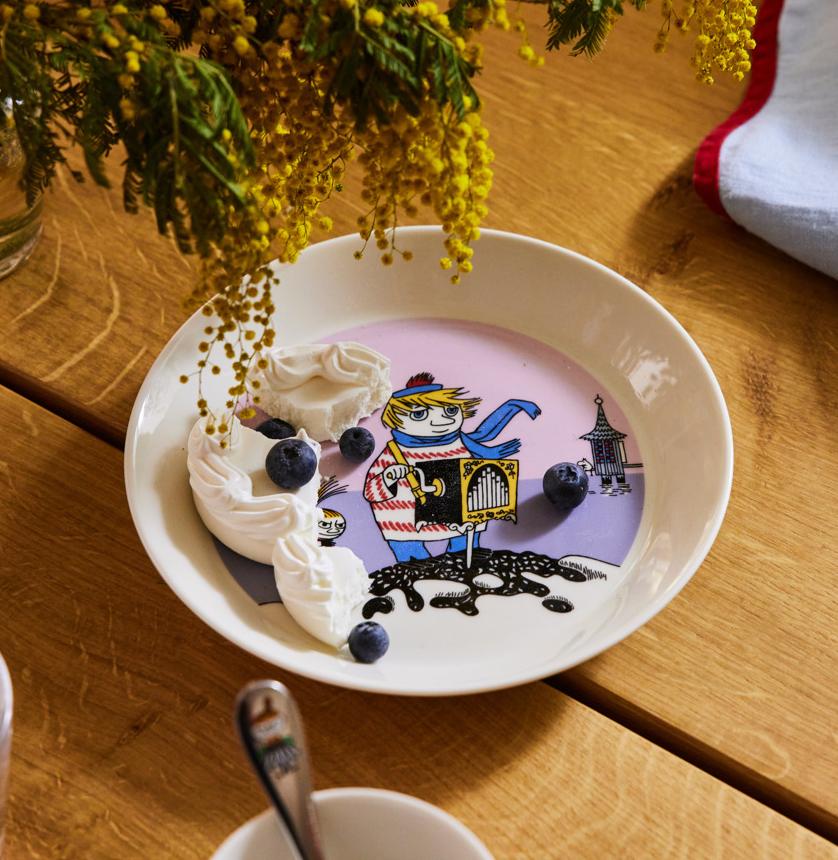En barnsligt illustrerad tallrik med vispad grädde och blåbär står på ett dukat träbord bredvid gula blommor.