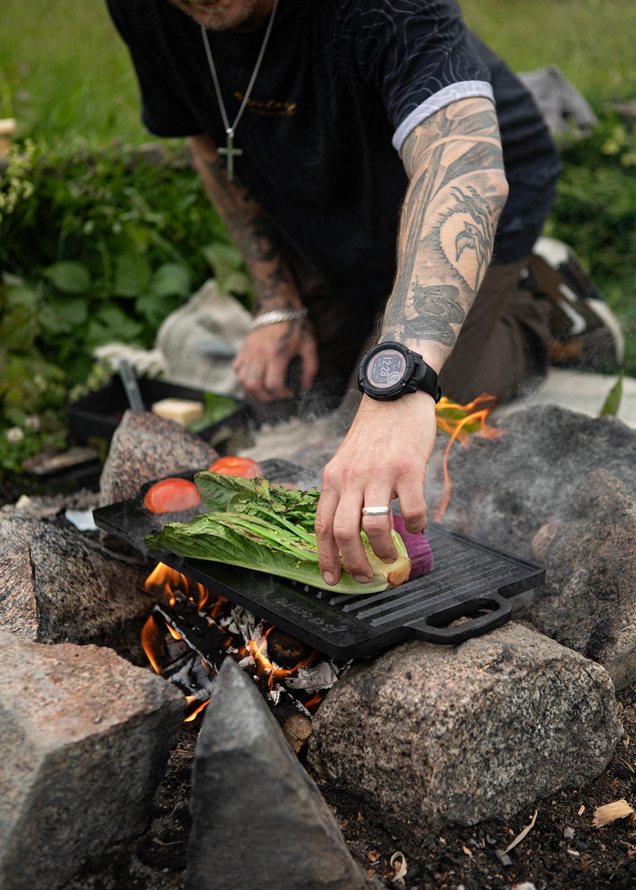 Person med tatueringar grillar färska grönsaker över öppen eld utomhus.