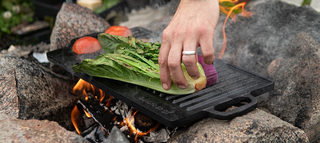 Tatuoitu henkilö grillaa vihanneksia avotulella luonnossa.