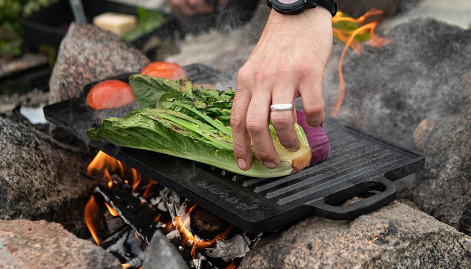 Tatuoitu henkilö grillaa vihanneksia avotulella luonnossa.