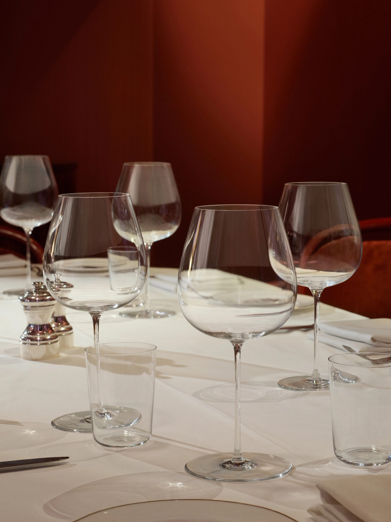 Ruokapöytä, jossa on valkoinen pöytäliina, useita viini- ja vesilaseja sekä suola- ja pippurisirotin lämpimänpunaista seinää vasten.